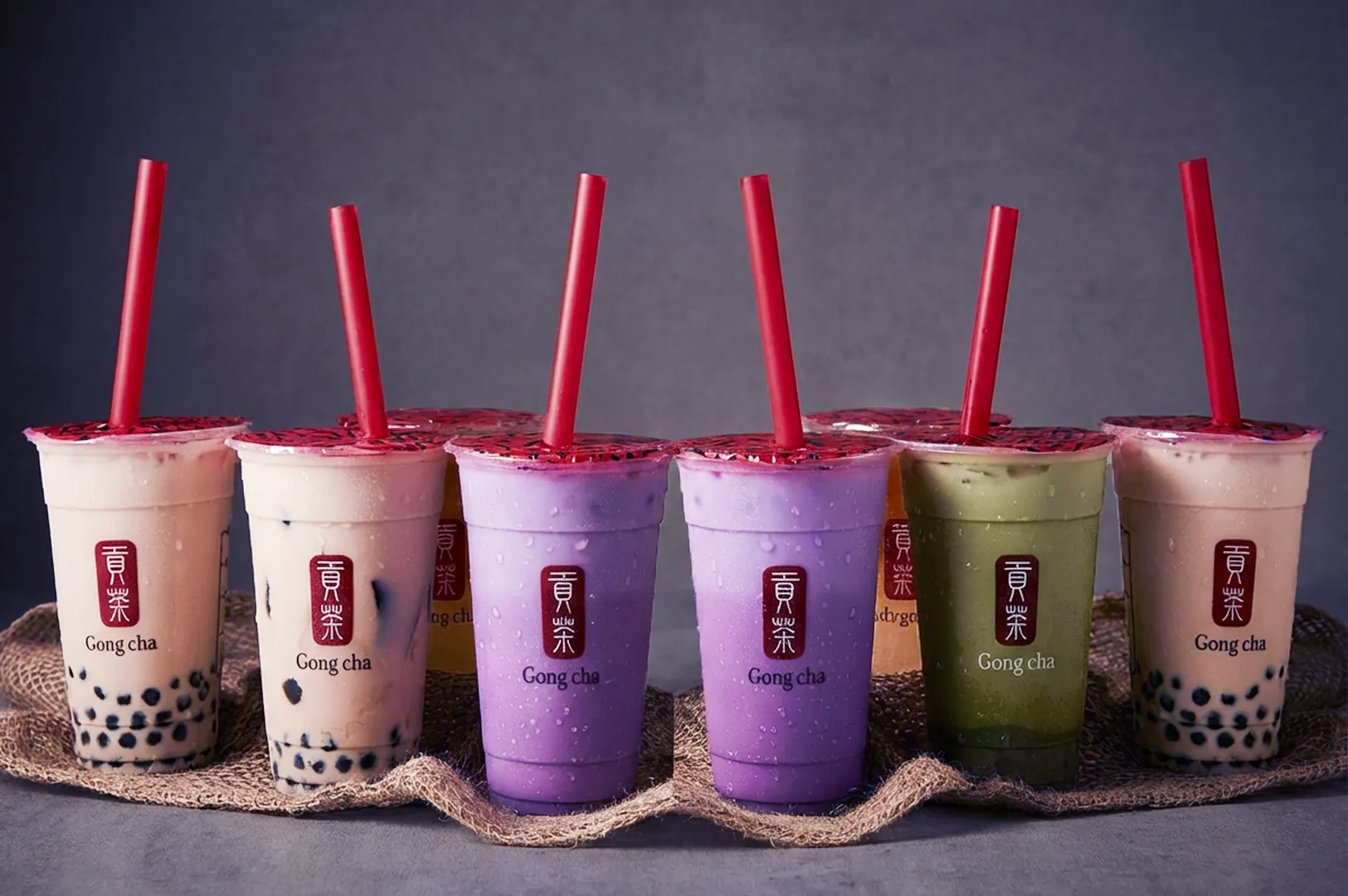 A row of bubble tea cups in various colors, including beige, purple, and green, with red straws on a textured cloth. The mood is inviting and vibrant.