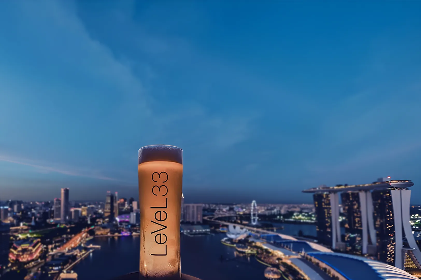 Tall glass with "Level33" logo against a vibrant evening cityscape. Bright lights illuminate the skyline under a serene blue sky, evoking a sense of luxury.
