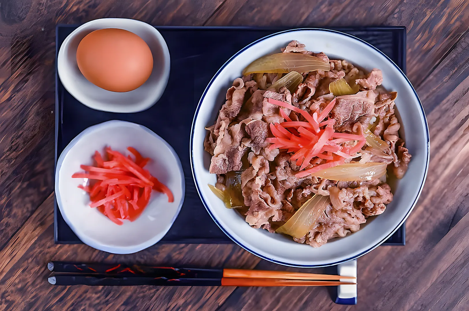 Bowl of gyudon with beef, onions, and pink pickled ginger on a dark tray. A raw egg and extra ginger are in small dishes beside it. Chopsticks rest nearby.