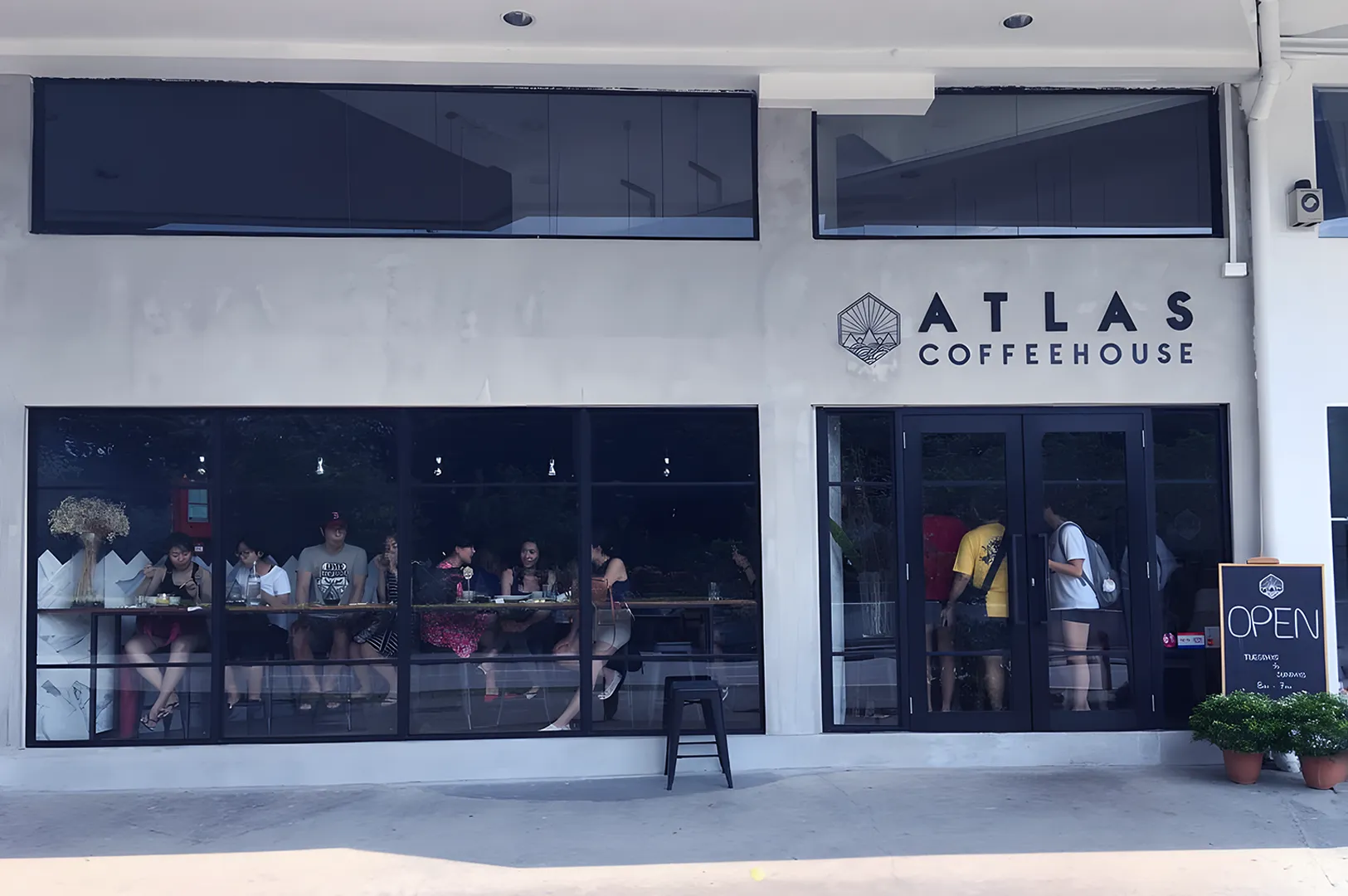 Atlas Coffeehouse exterior with large windows showing patrons at tables inside. The facade is light grey with a logo and "OPEN" sign outside, conveying a welcoming vibe.