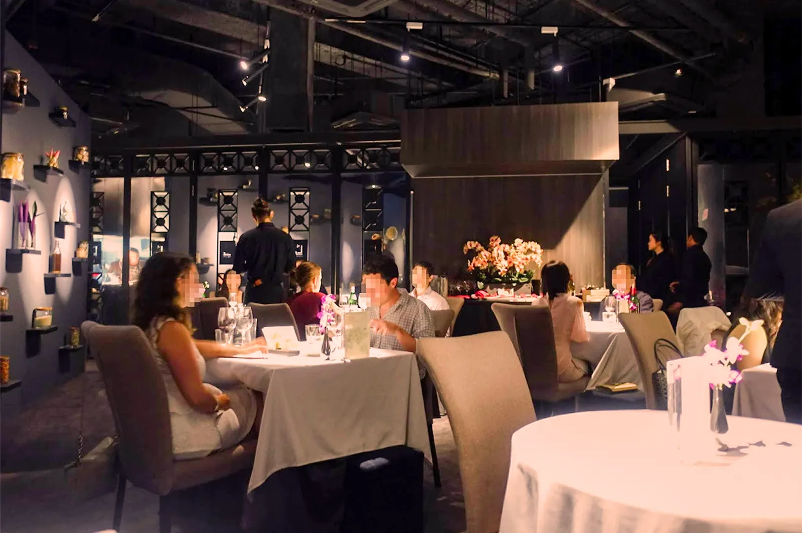 Dimly lit restaurant with elegant decor, featuring tables set with white cloths and vases of flowers. Patrons enjoy a sophisticated dining experience.