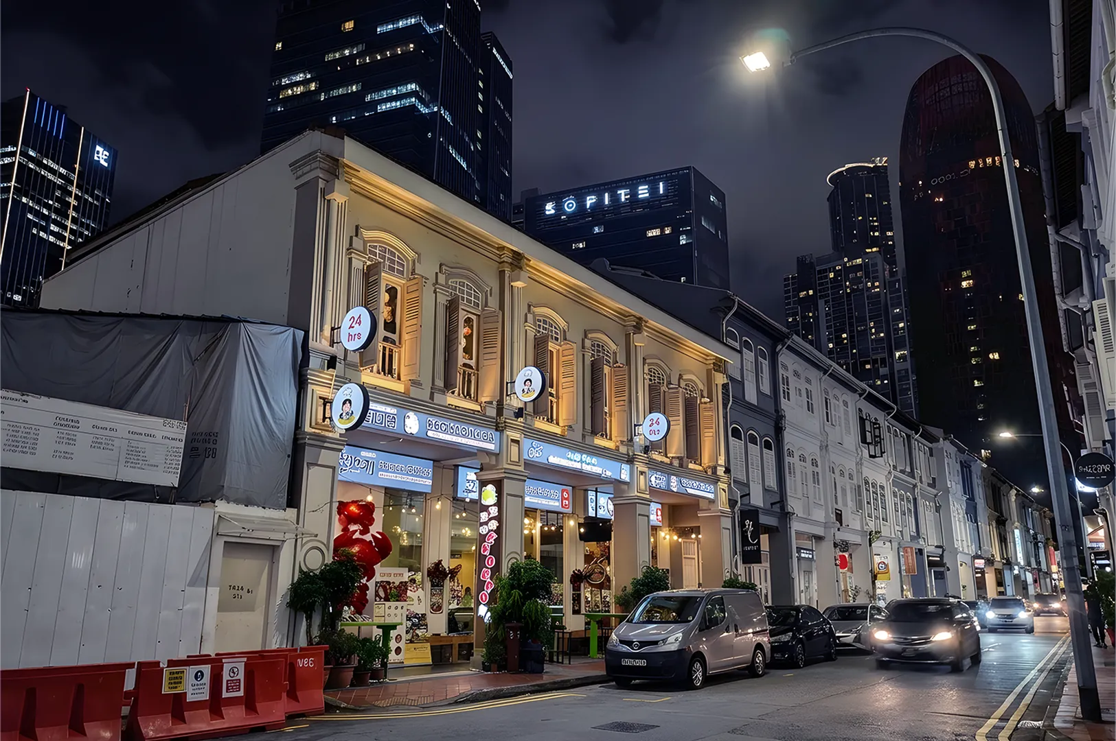 Illuminated street at night with historic shophouses featuring bright storefronts. Modern skyscrapers loom in the background under a cloudy sky. Cars pass by, adding a lively urban feel.