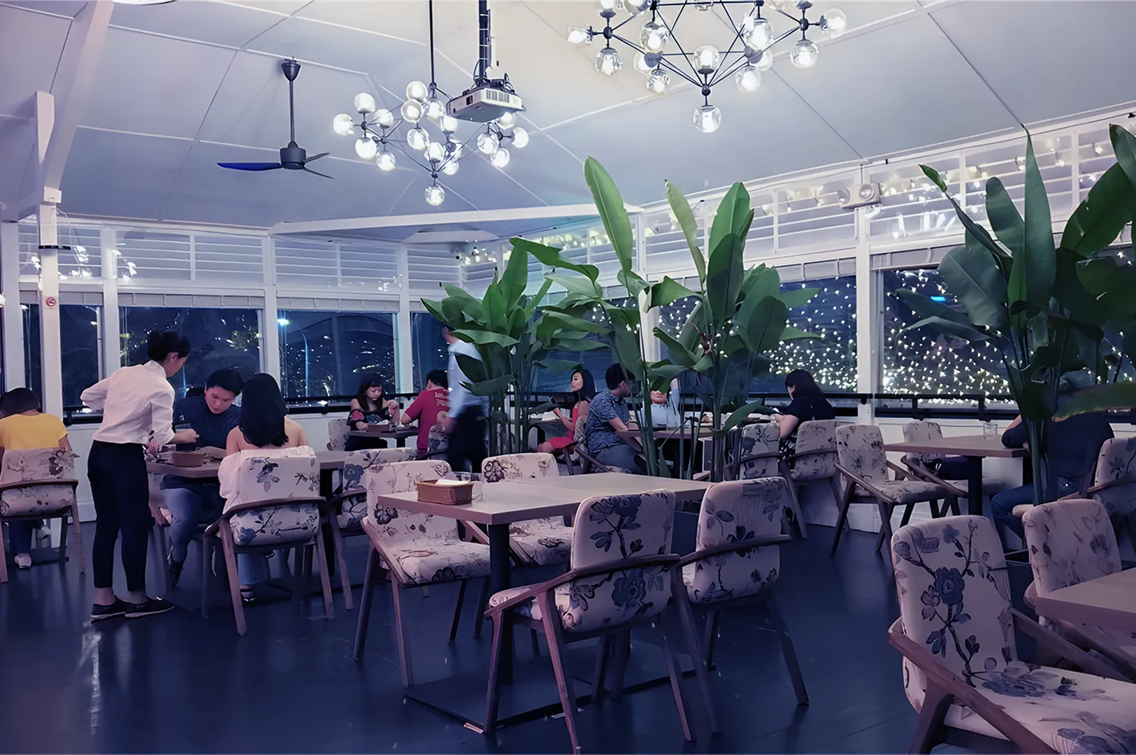 Modern restaurant interior with people dining, featuring floral chairs, wooden tables, decorative plants, and elegant chandeliers, creating a cozy ambiance.