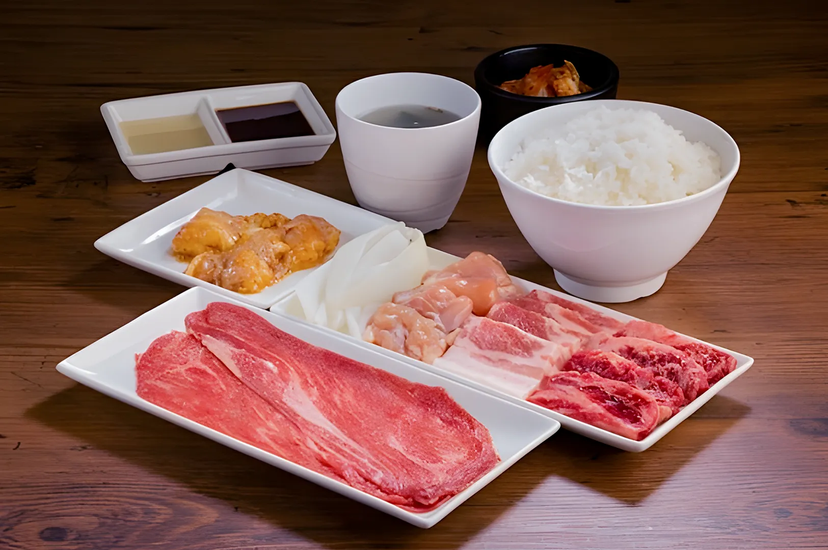 A wooden table with a three-part white tray of sliced meats and seafood, a bowl of white rice, small bowls of sauces, and kimchi, creating a traditional and appetizing setting.