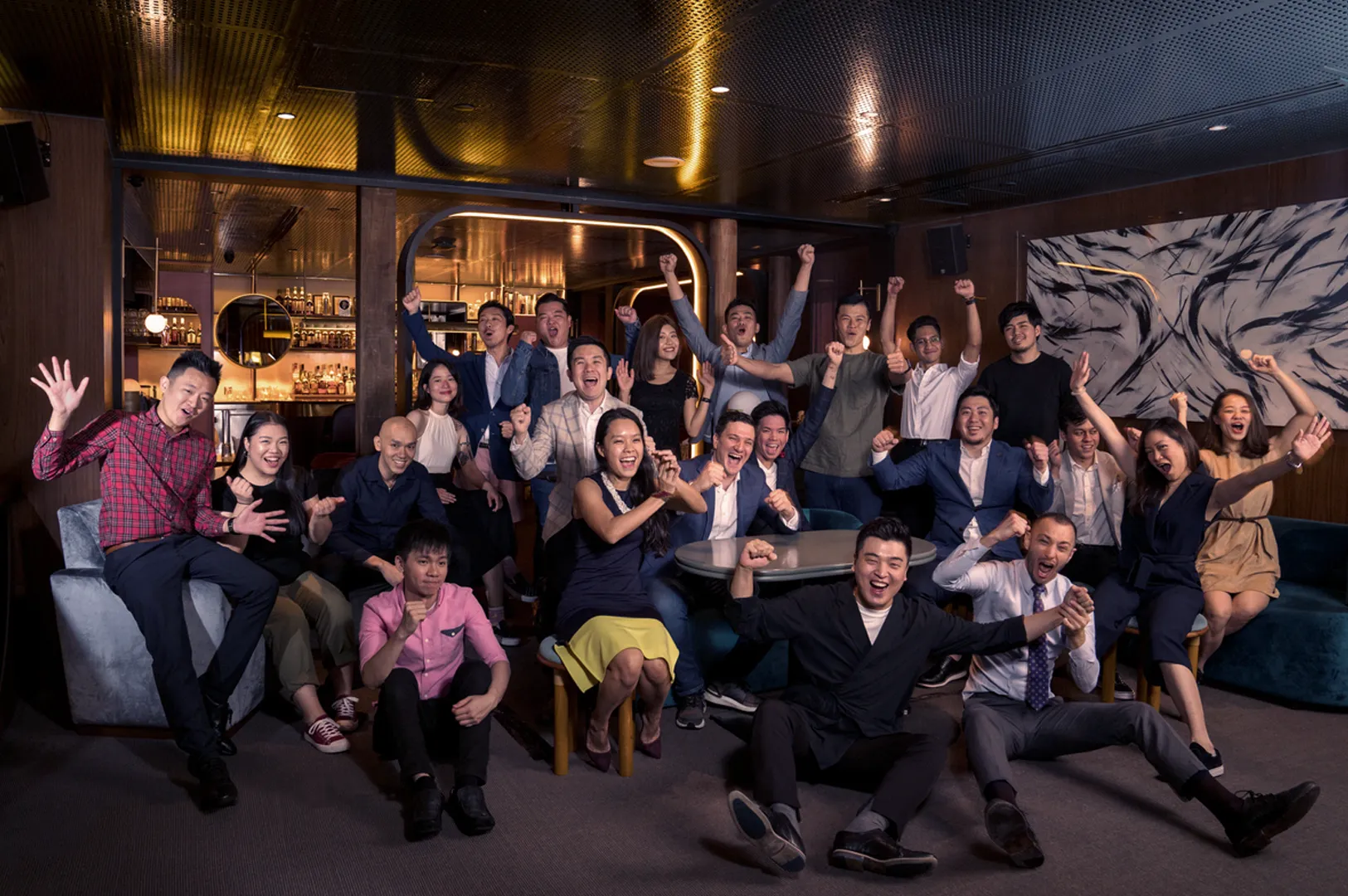 A diverse group of people joyfully cheering and posing in a dimly lit, stylish bar. The atmosphere is lively and celebratory, with smiling faces and raised arms.