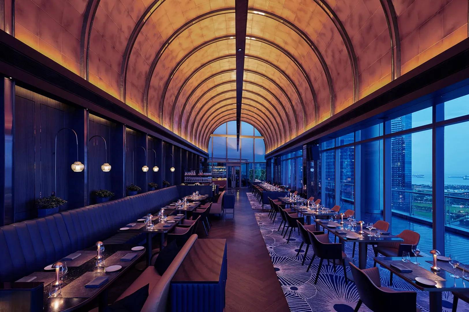 Elegant restaurant with arched ceiling and ambient lighting, featuring intimate table settings and large windows showing a cityscape at dusk.