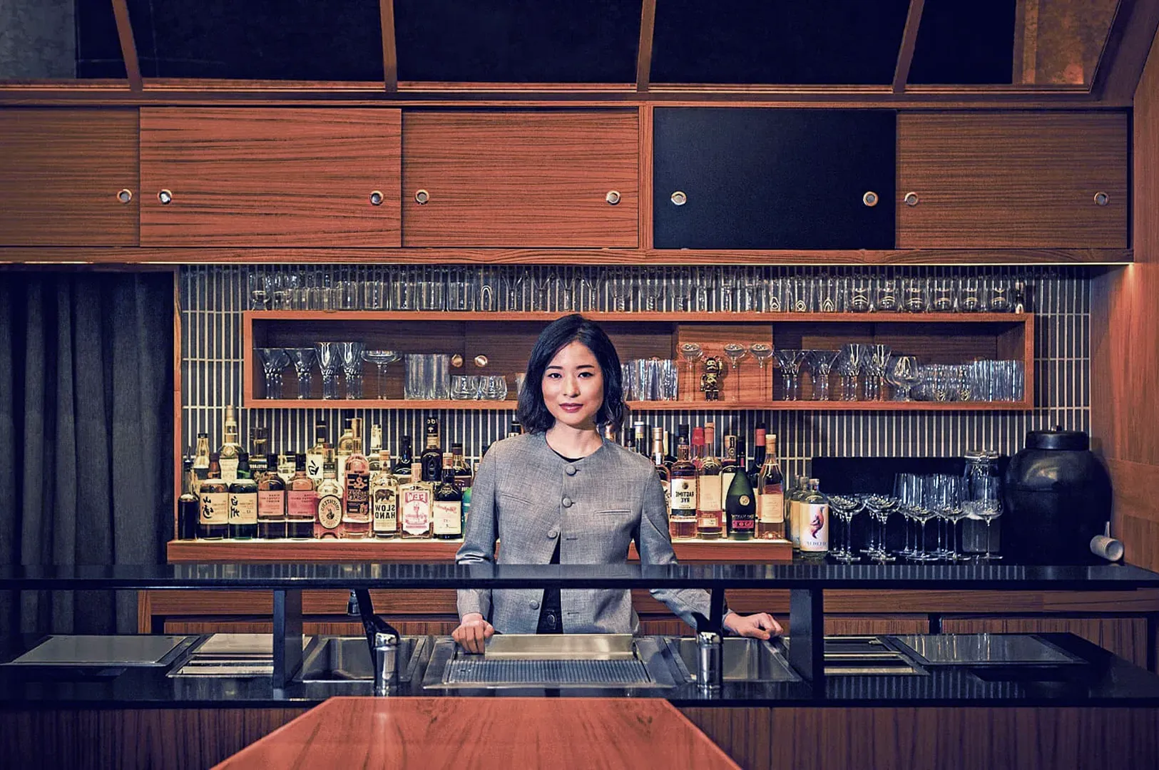 A woman stands confidently behind a well-stocked bar, surrounded by shelves of glasses and liquor bottles, conveying a refined and professional atmosphere.
