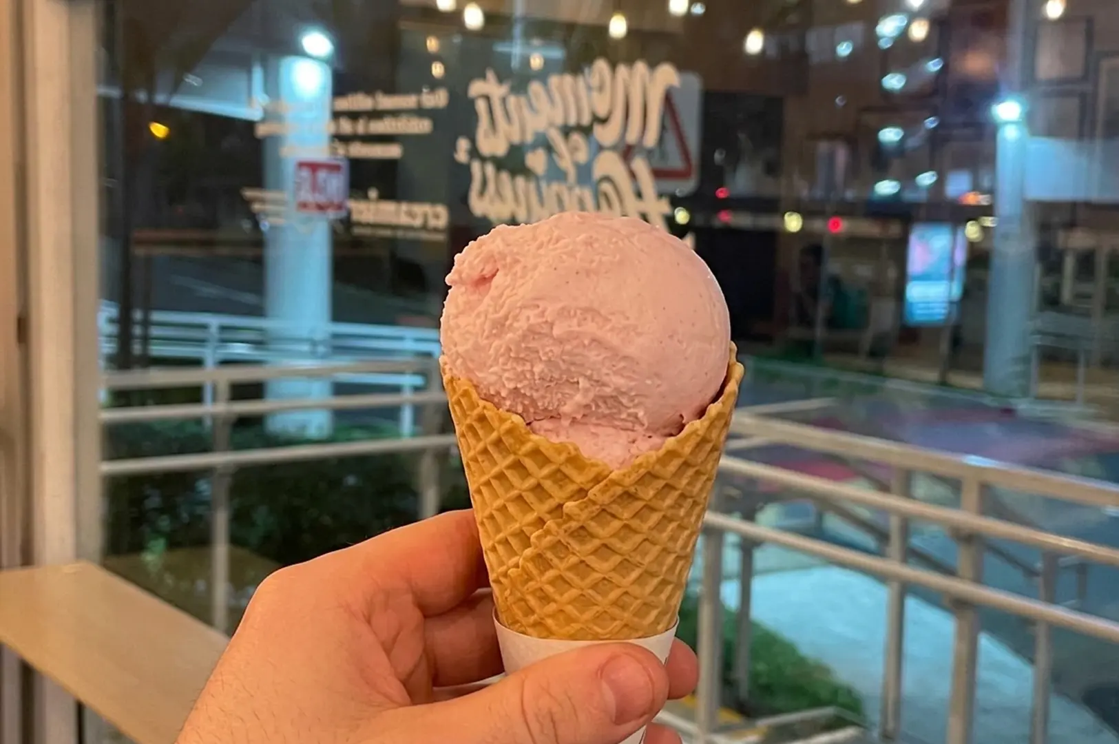 Hand holding a pink ice cream scoop in a waffle cone in front of a shop window at night