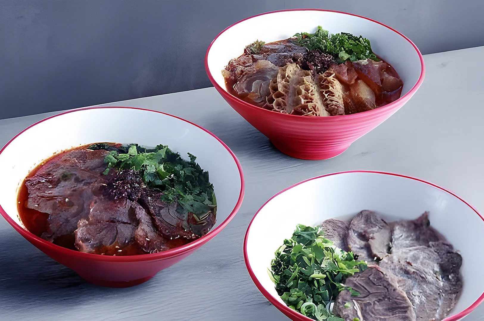 Three red-rimmed bowls of Chinese noodle soup on a gray table. Each bowl has different meats and herbs, creating a warm, savory atmosphere.