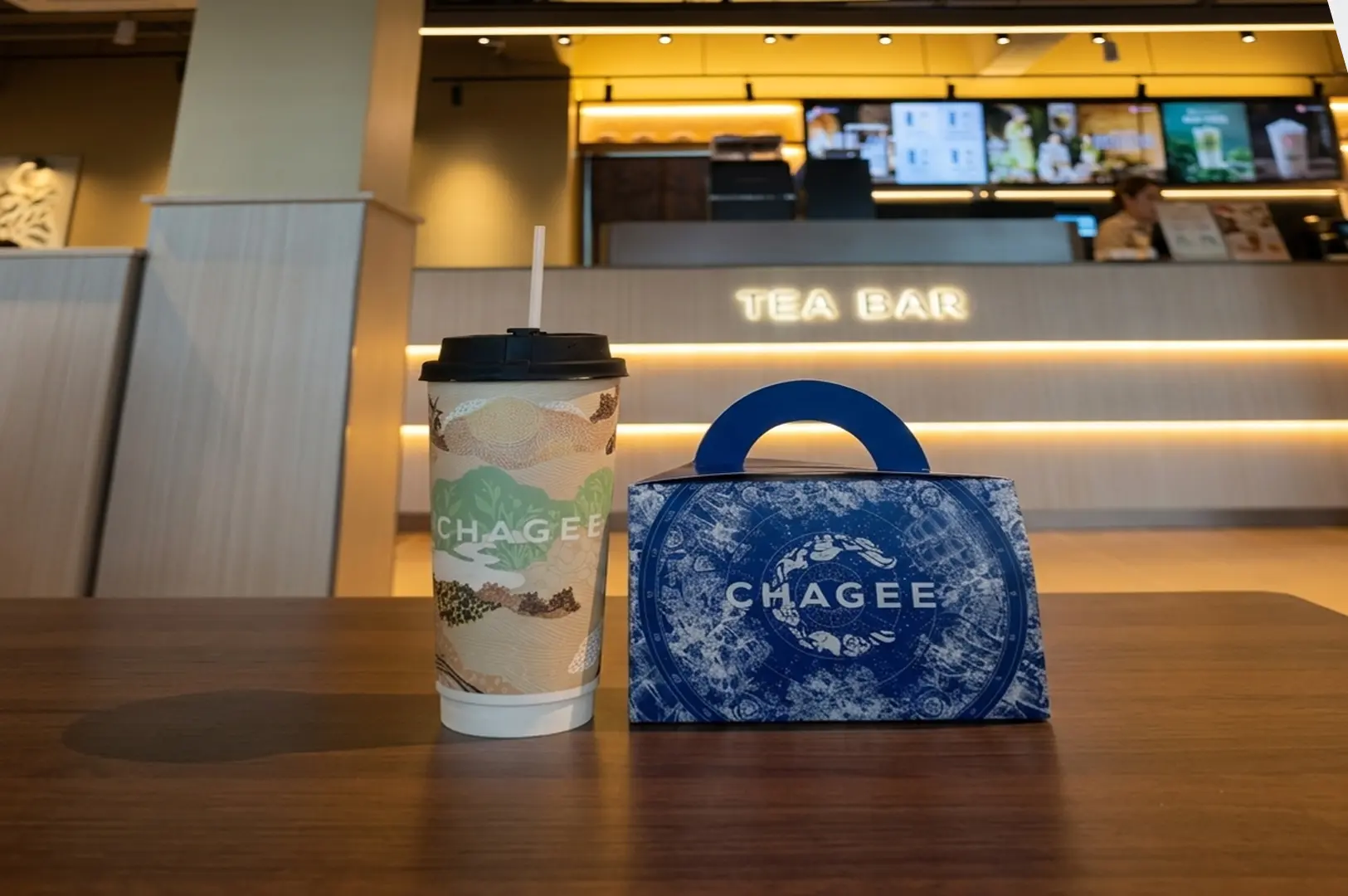 Chagee branded tea cup and blue decorative carrying box displayed on wooden table at modern tea bar with warm lighting and illuminated signage