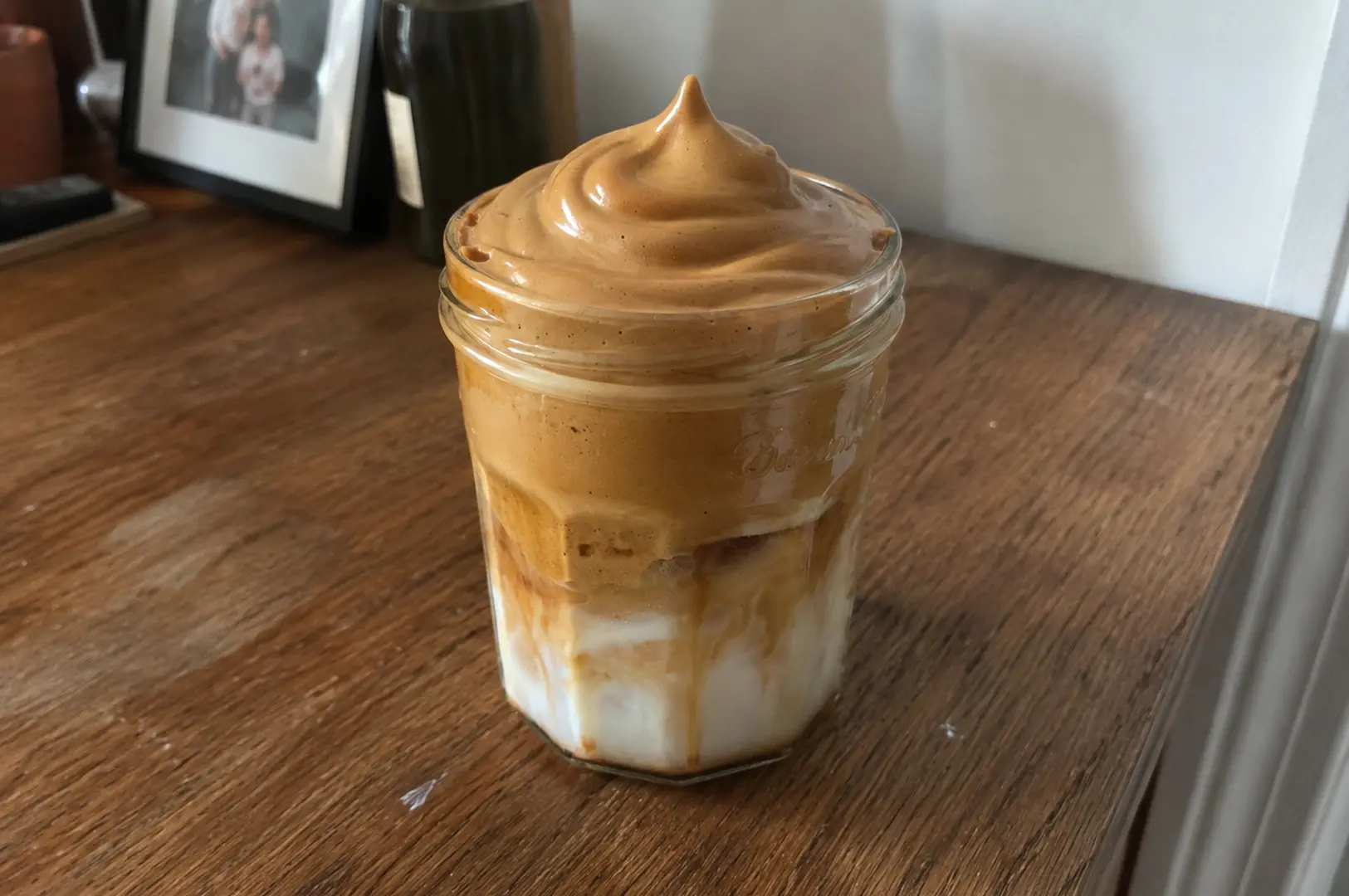 Glass jar of Dalgona coffee featuring thick whipped coffee foam over milk and ice on a wooden surface