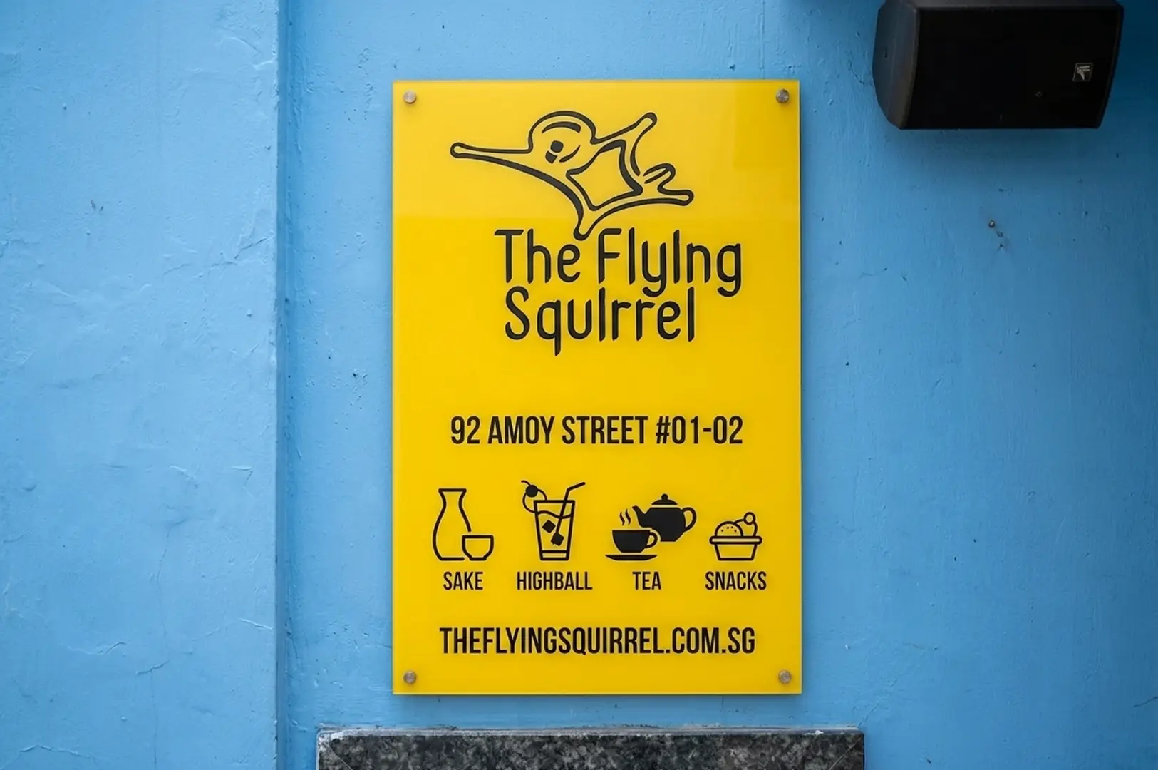 Bright yellow sign on a blue wall displaying The Flying Squirrel logo, address at 92 Amoy Street, and icons for sake, highball, tea, and snacks