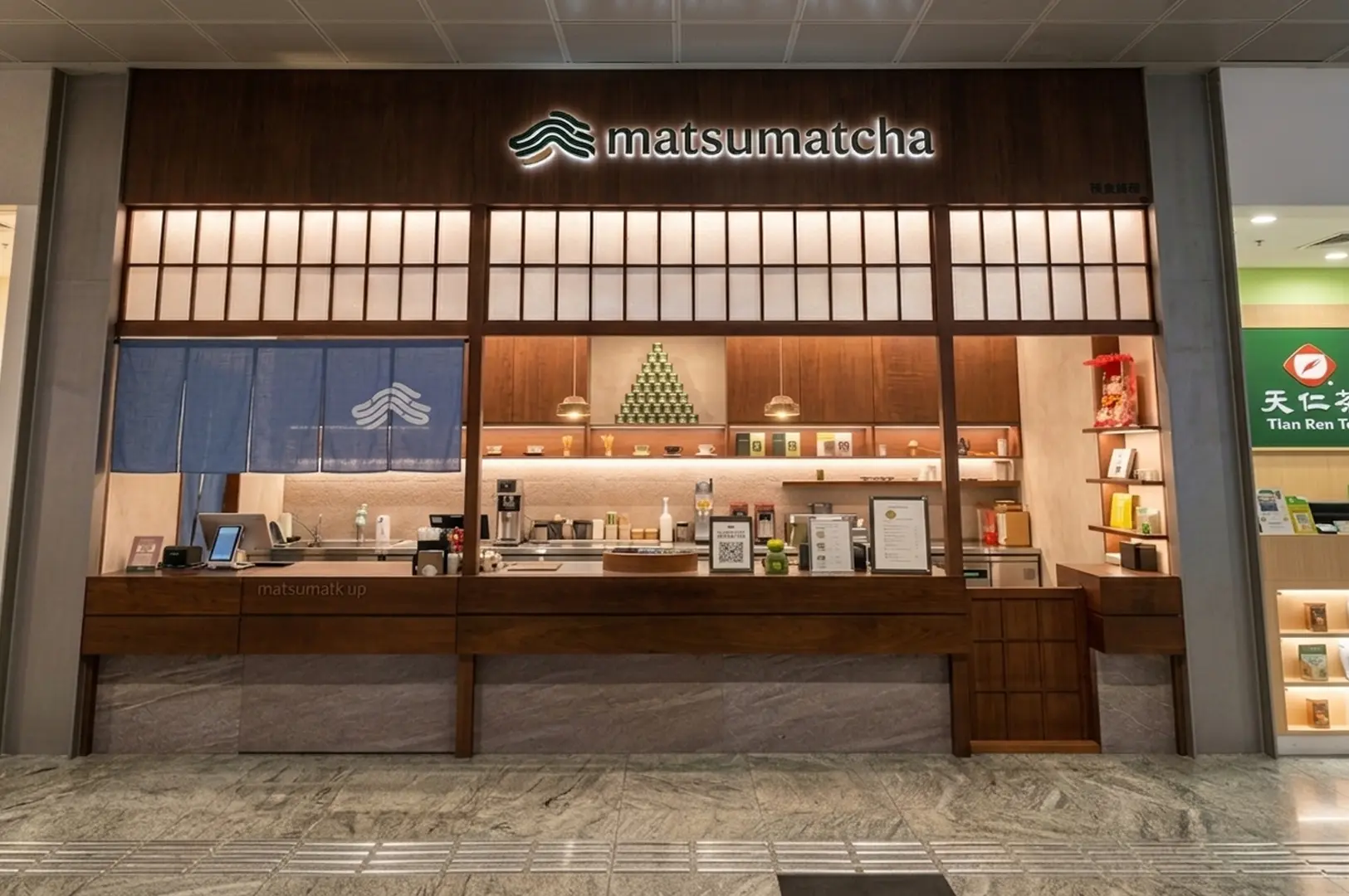 Storefront of matsumatcha tea shop featuring a wooden counter, shoji-style panels, and a blue noren curtain with the brand logo
