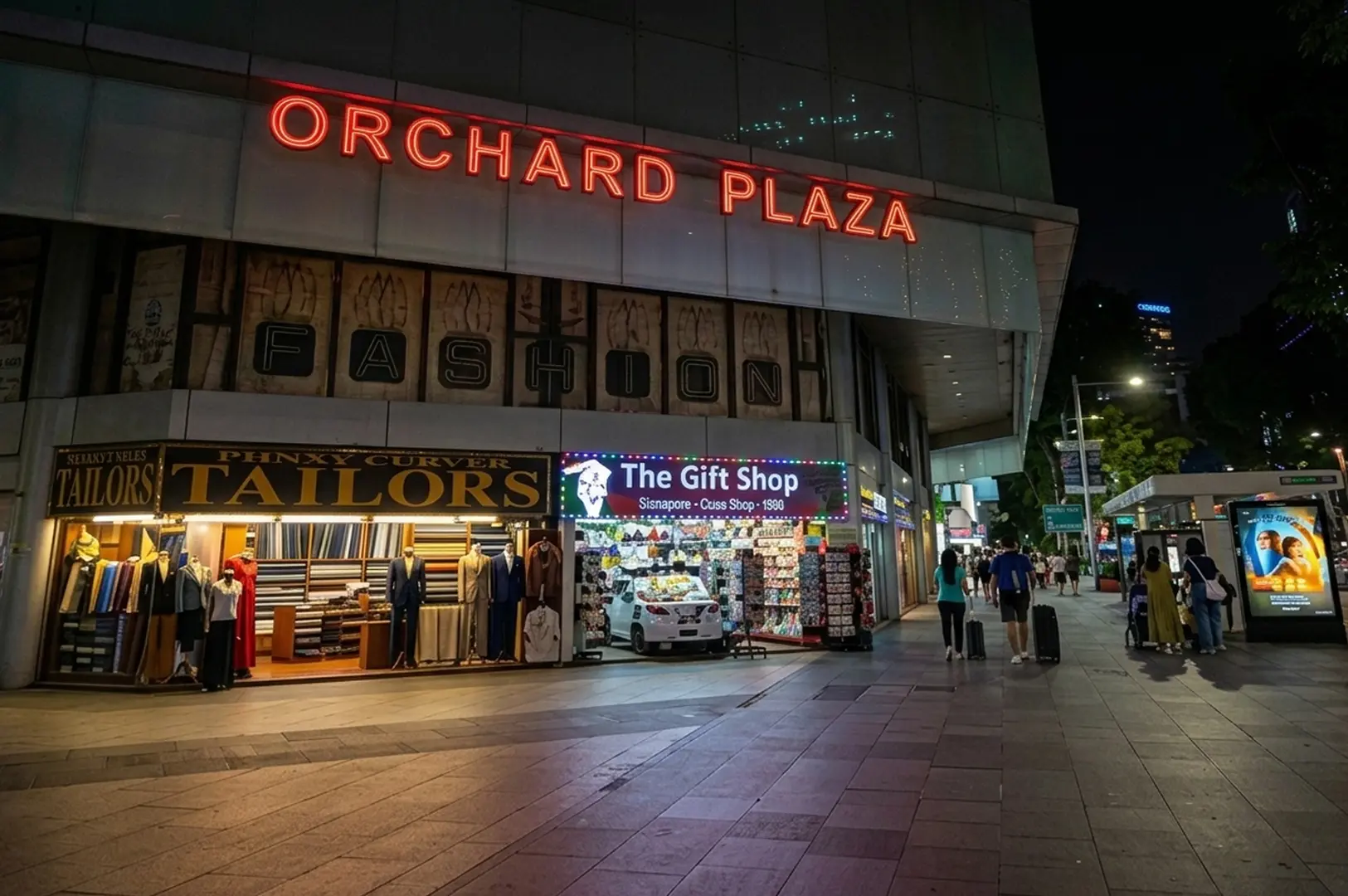 Night view of Orchard Plaza's illuminated neon sign with street-level shops including tailors and gift stores