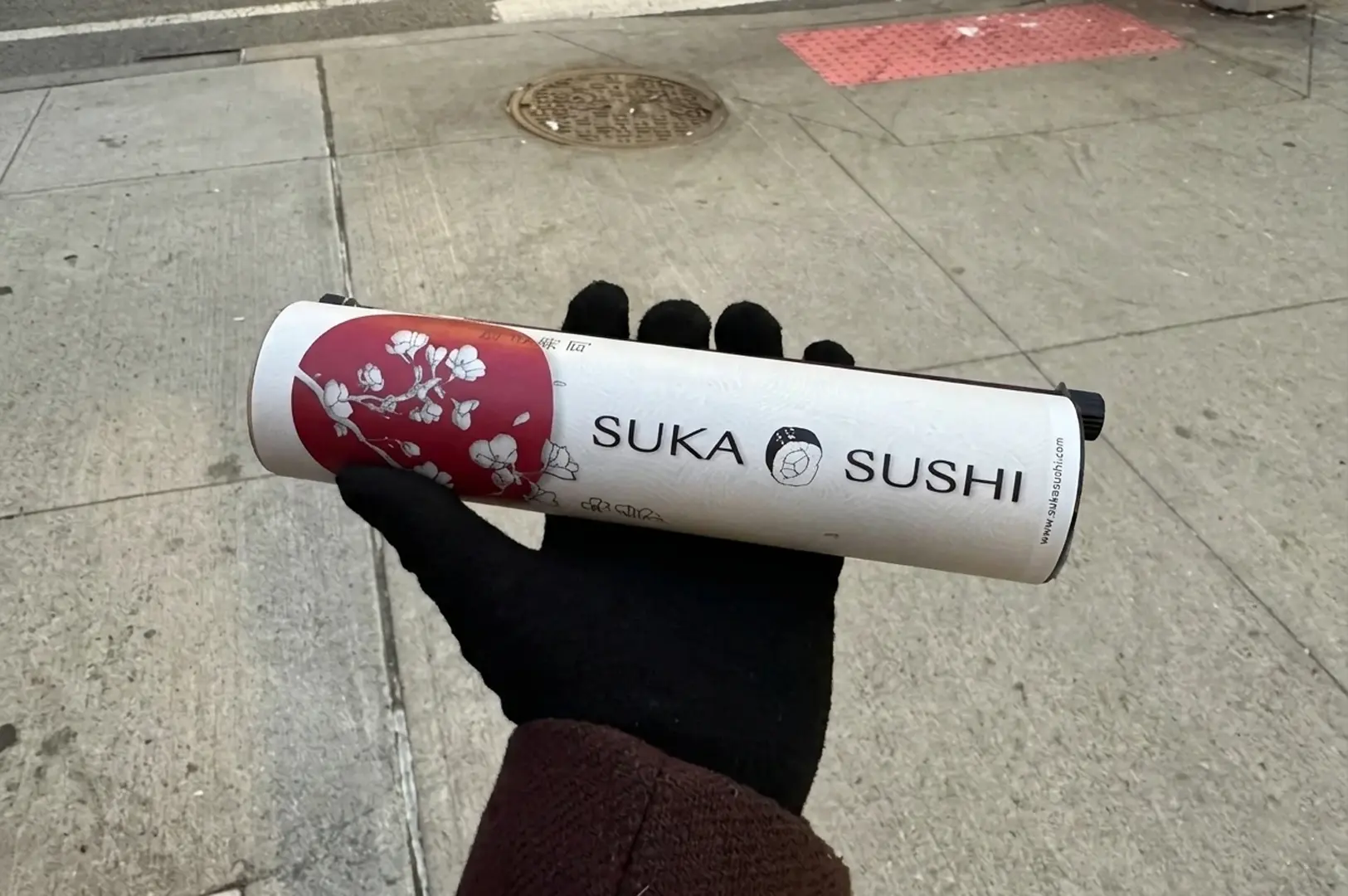 Hand in black glove holding a white cylindrical tube labeled Suka Sushi with red cherry blossom design on a sidewalk pavement