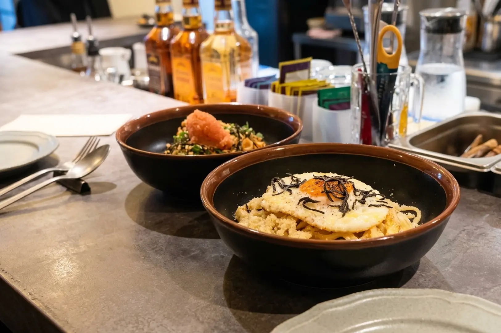 Two brown ceramic bowls of Japanese-style rice dishes on a restaurant counter with condiments and bottles in background