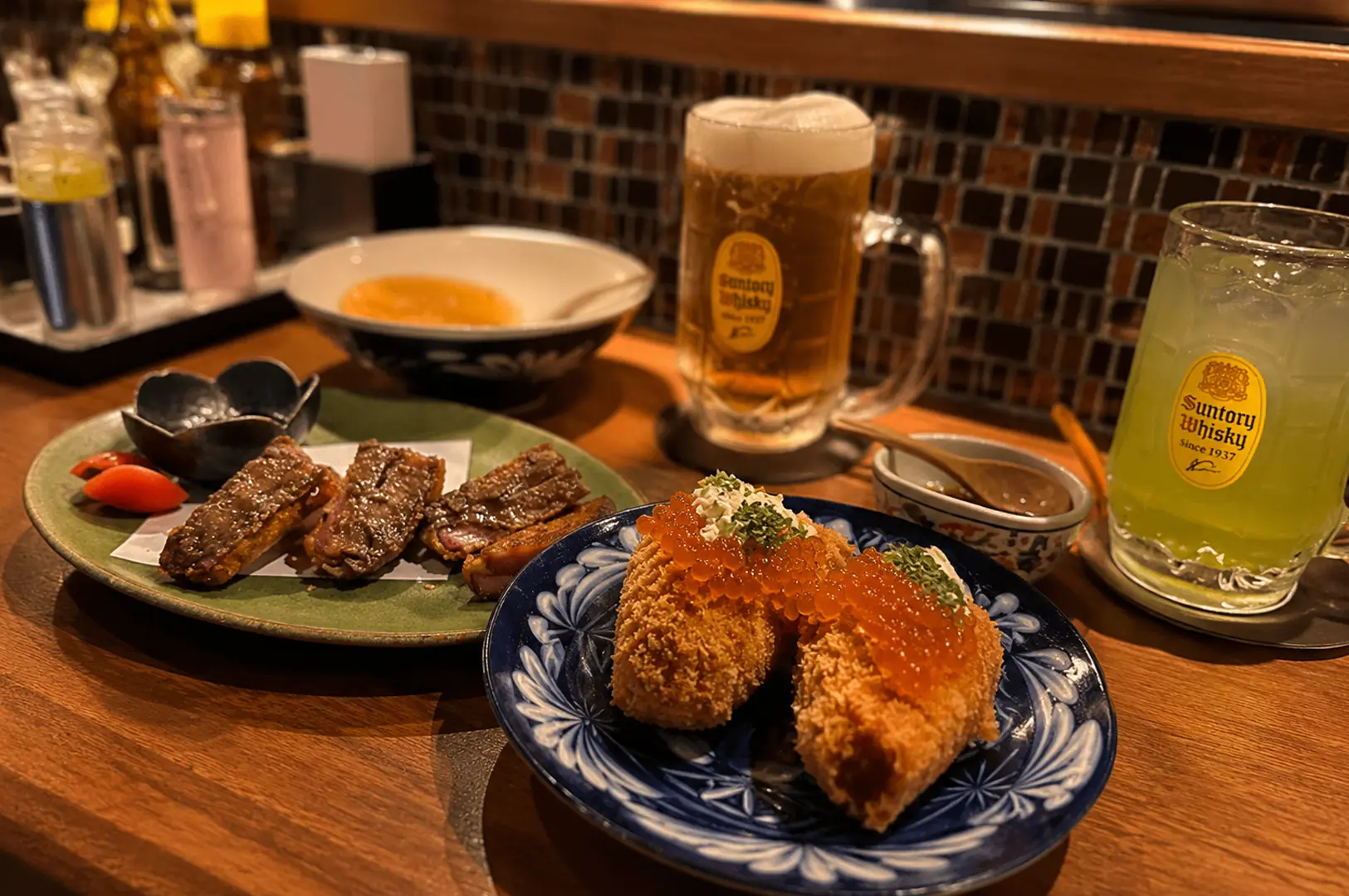 Japanese izakaya feast featuring croquettes with salmon roe, grilled beef, drinks, and side dishes on a wooden counter.