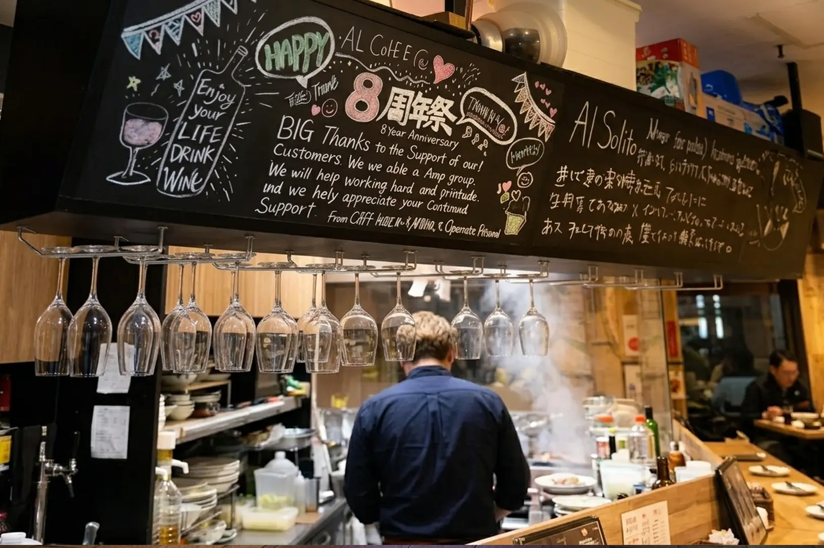 Interior view of Al Solito restaurant featuring a chalkboard sign celebrating its 8th anniversary, hanging wine glasses, and a chef working in the kitchen