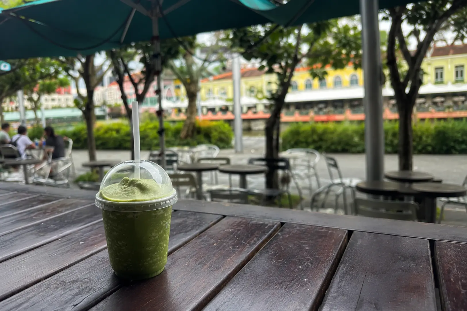 Iced matcha drink in plastic cup with dome lid on outdoor wooden table at Clarke Quay with colorful riverside buildings and green umbrellas in background