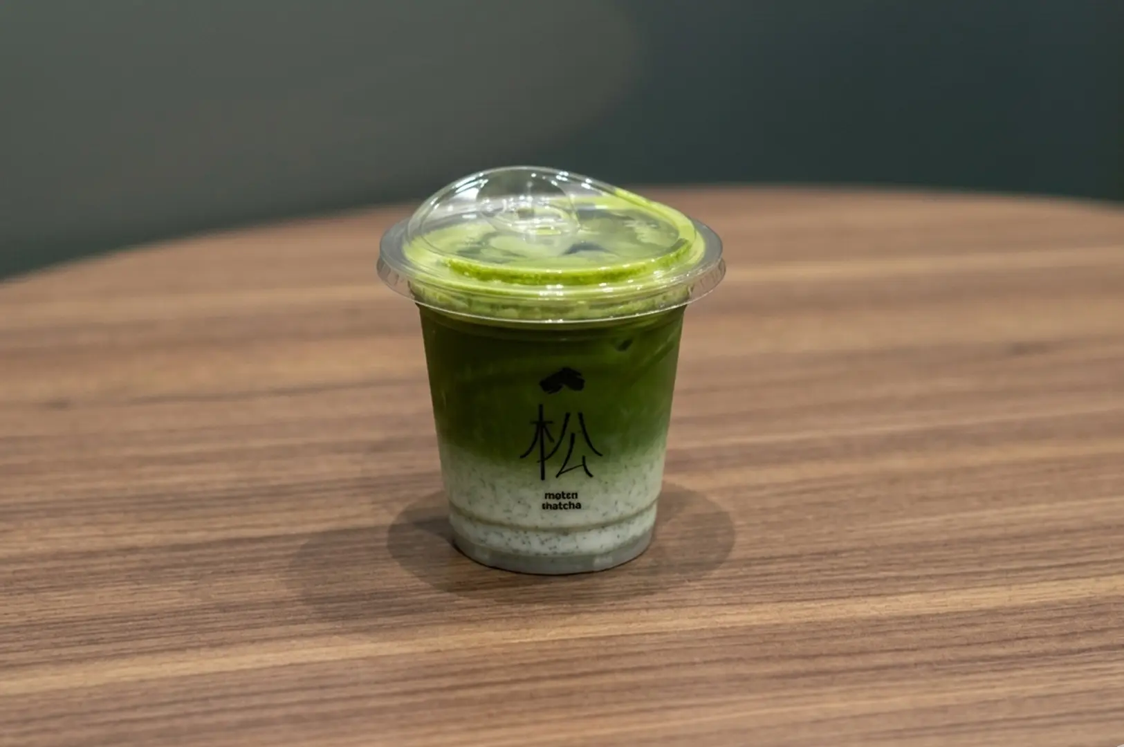 Layered iced matcha latte in a clear plastic cup with the matsumatcha logo sitting on a wooden table