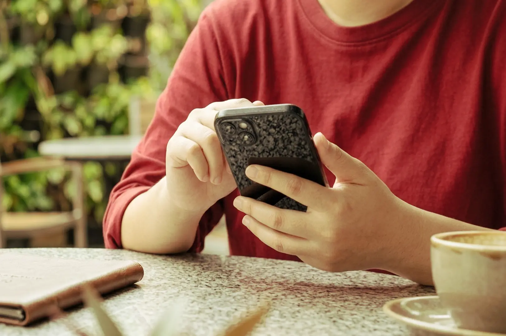 Person in red shirt scrolling phone at cafe table with coffee