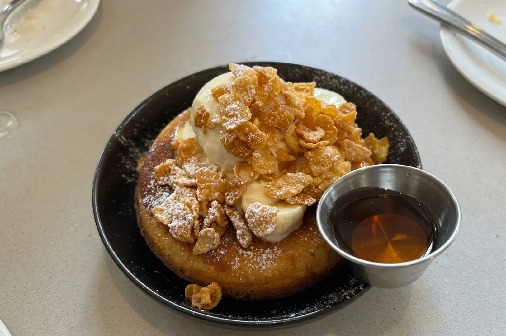 Fluffy pancake topped with a scoop of ice cream, cornflakes, and powdered sugar, served with a side of syrup on a black plate