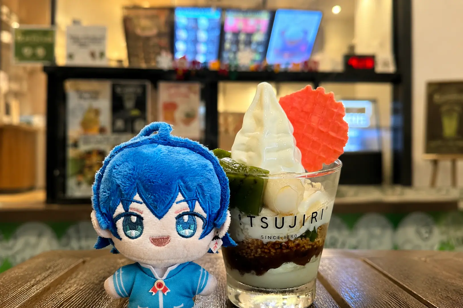 Blue-haired plush toy character sitting beside TSUJIRI parfait dessert with soft serve ice cream and wafer on wooden table inside the cafe