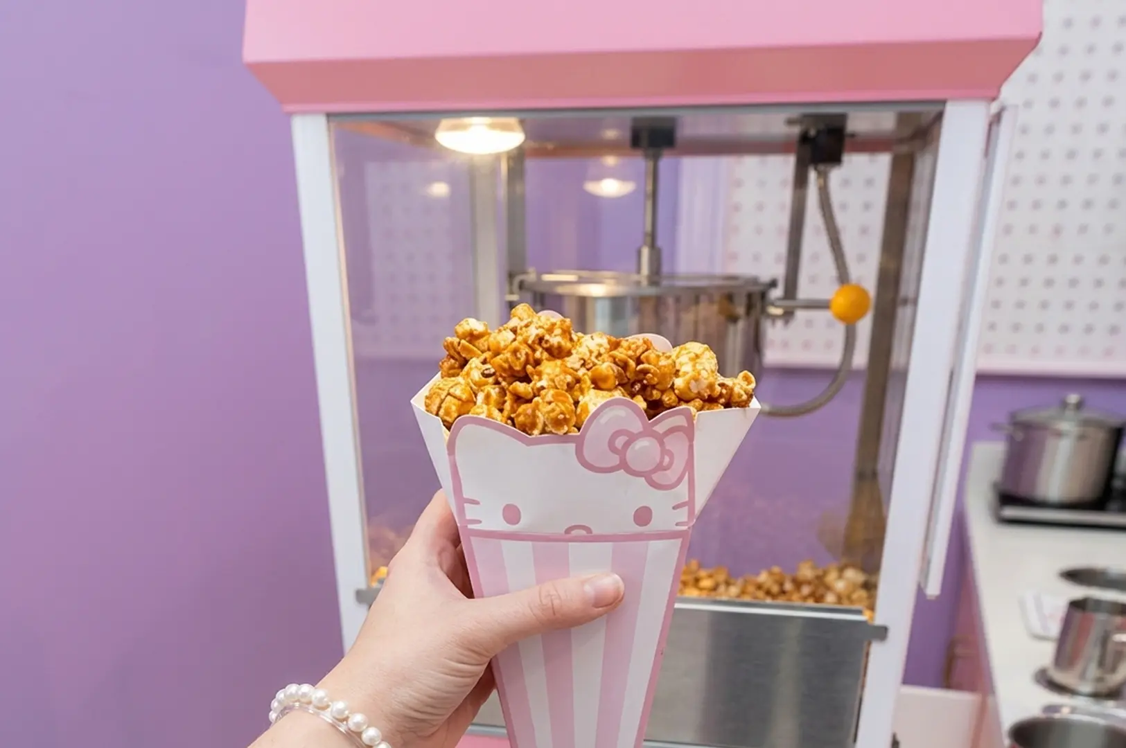 Hand holding Hello Kitty branded popcorn cone filled with caramel popcorn in front of pink popcorn machine at Universal Studios Singapore