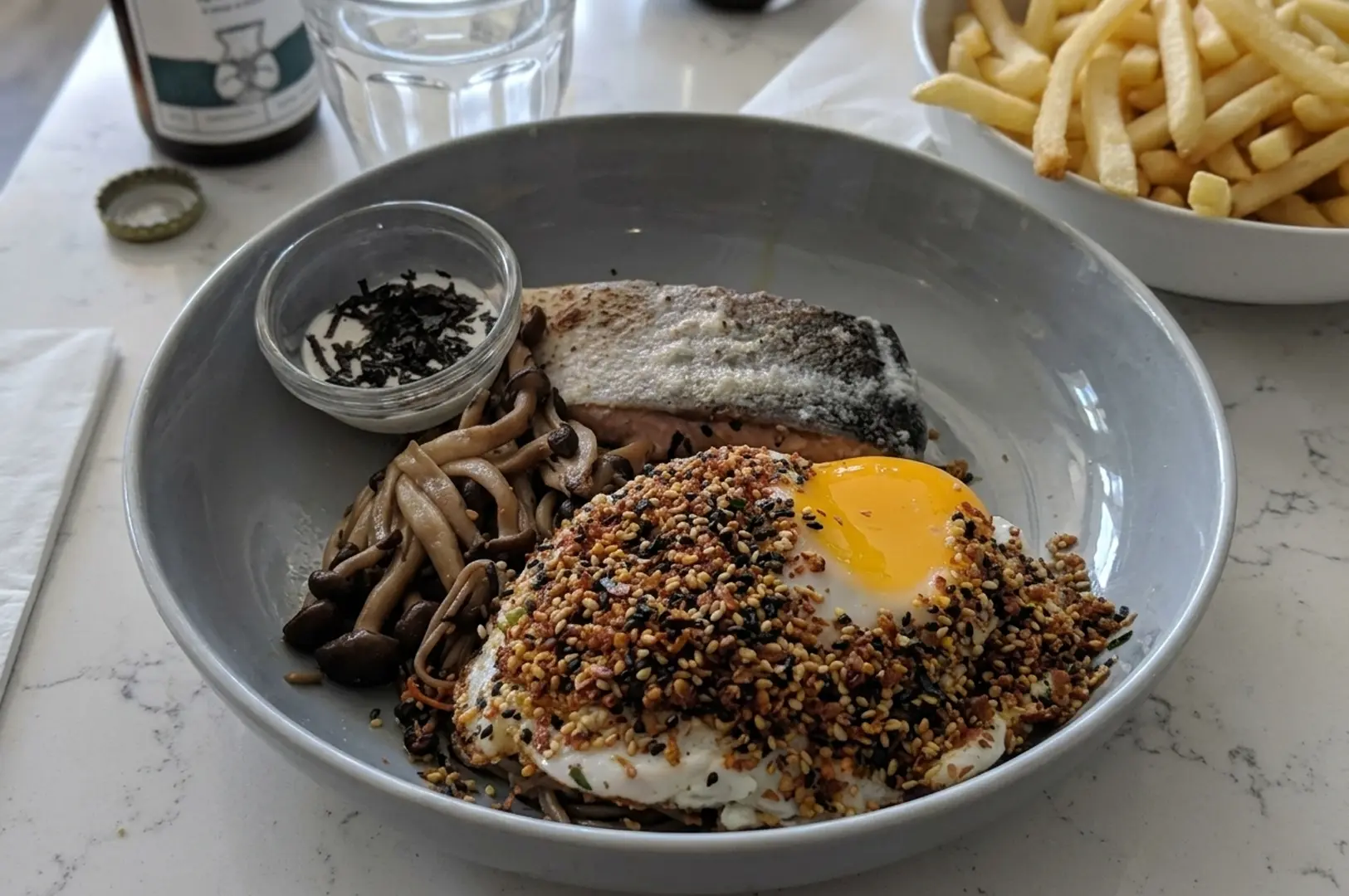 Savory bowl featuring seared salmon, sautéed mushrooms, and a fried egg topped with furikake seasoning, served with a side of french fries