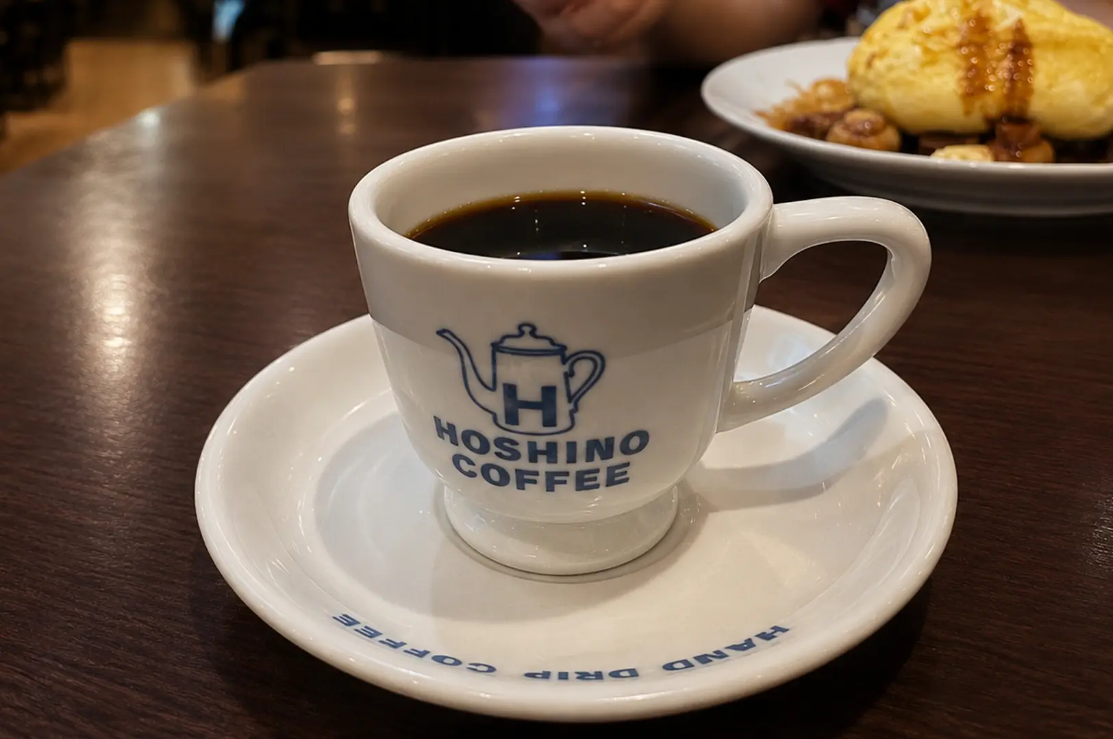 White ceramic cup of black coffee with the blue Hoshino Coffee logo on a saucer, with a plate of food in the background