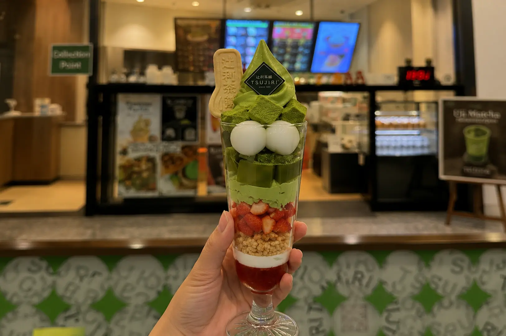 Hand holding tall matcha parfait glass with layers of strawberry, cereal, mochi balls, matcha cream, and soft serve ice cream topped with TSUJIRI wafer cookie at cafe counter