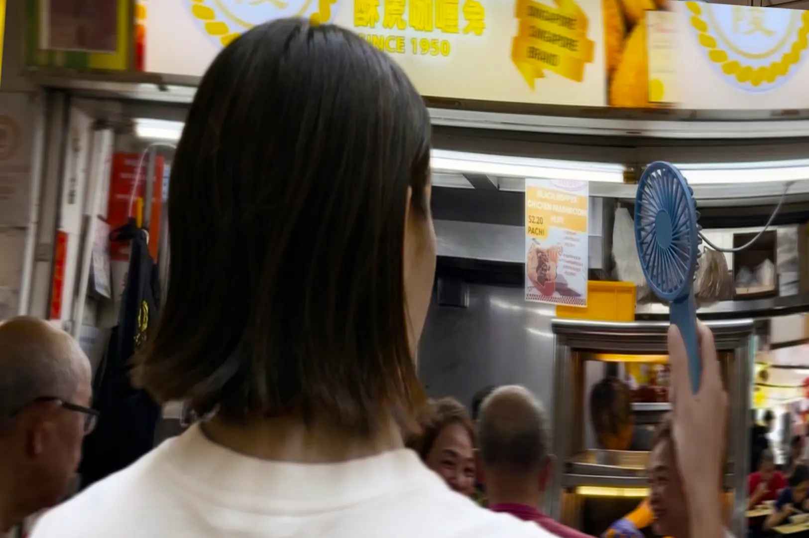 Person holding blue portable fan while waiting at crowded hawker center food stall