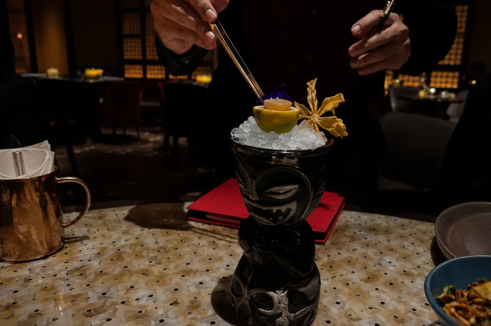 Bartender garnishing a tiki cocktail in a black face mug with crushed ice and flowers on a mother-of-pearl table