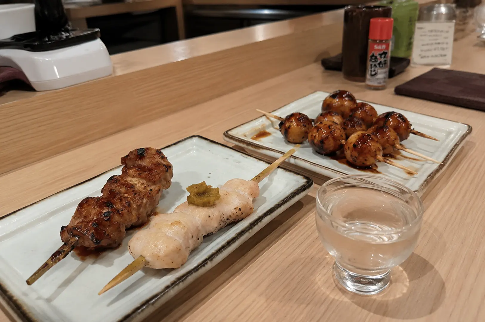 Assorted yakitori skewers with grilled chicken meatballs and a glass of sake at a traditional Japanese counter.