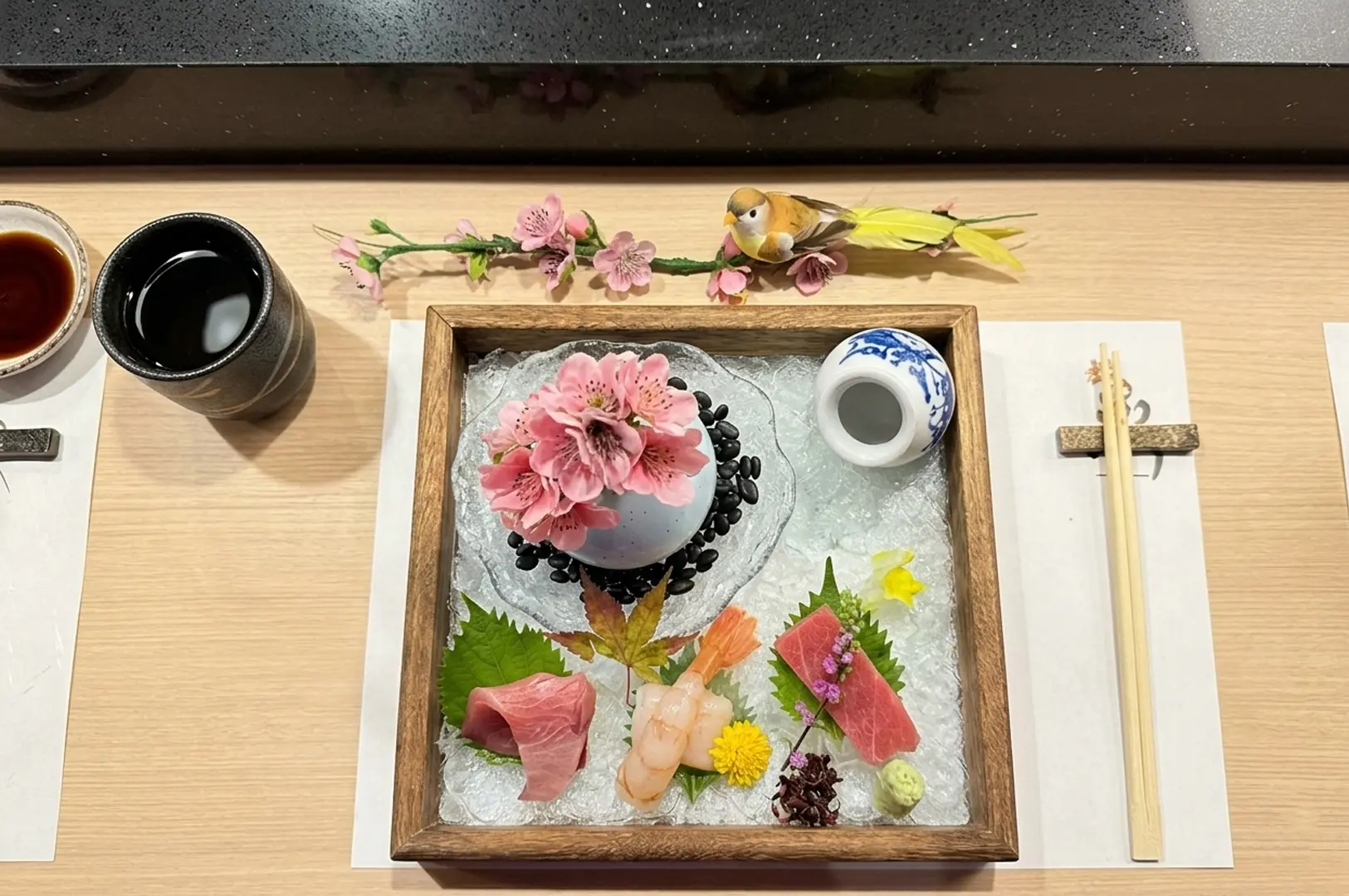 Elegant sashimi box presentation on ice featuring fresh fish slices, pink flowers, and a decorative bird figurine