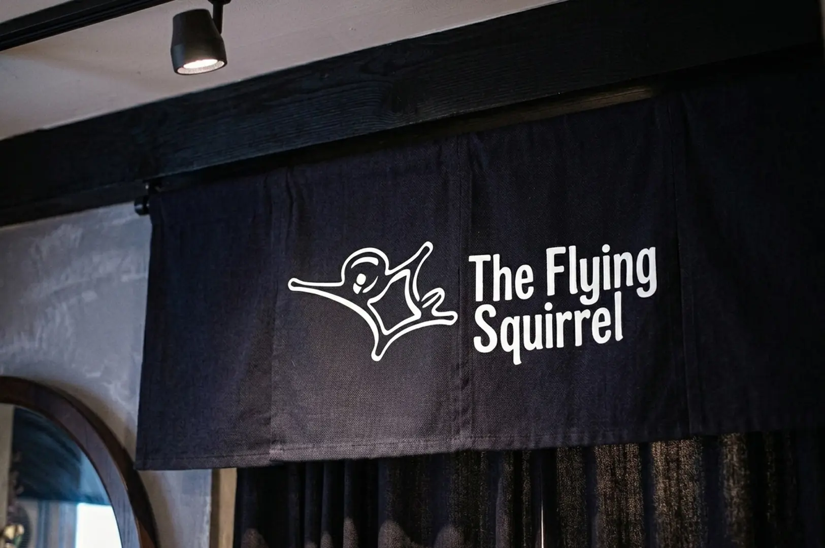 Black fabric noren curtain featuring the white Flying Squirrel logo and name hanging above a doorway