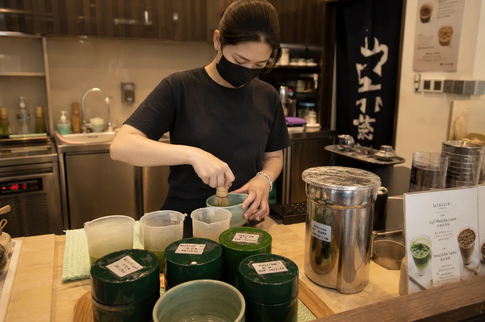 Staff member whisking fresh matcha tea in traditional bowl behind counter with green tea canisters