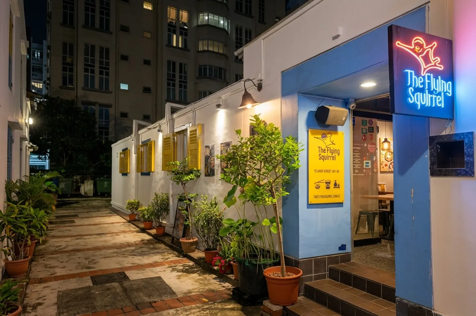 Night view of The Flying Squirrel restaurant exterior with blue walls, neon sign, and yellow shutters