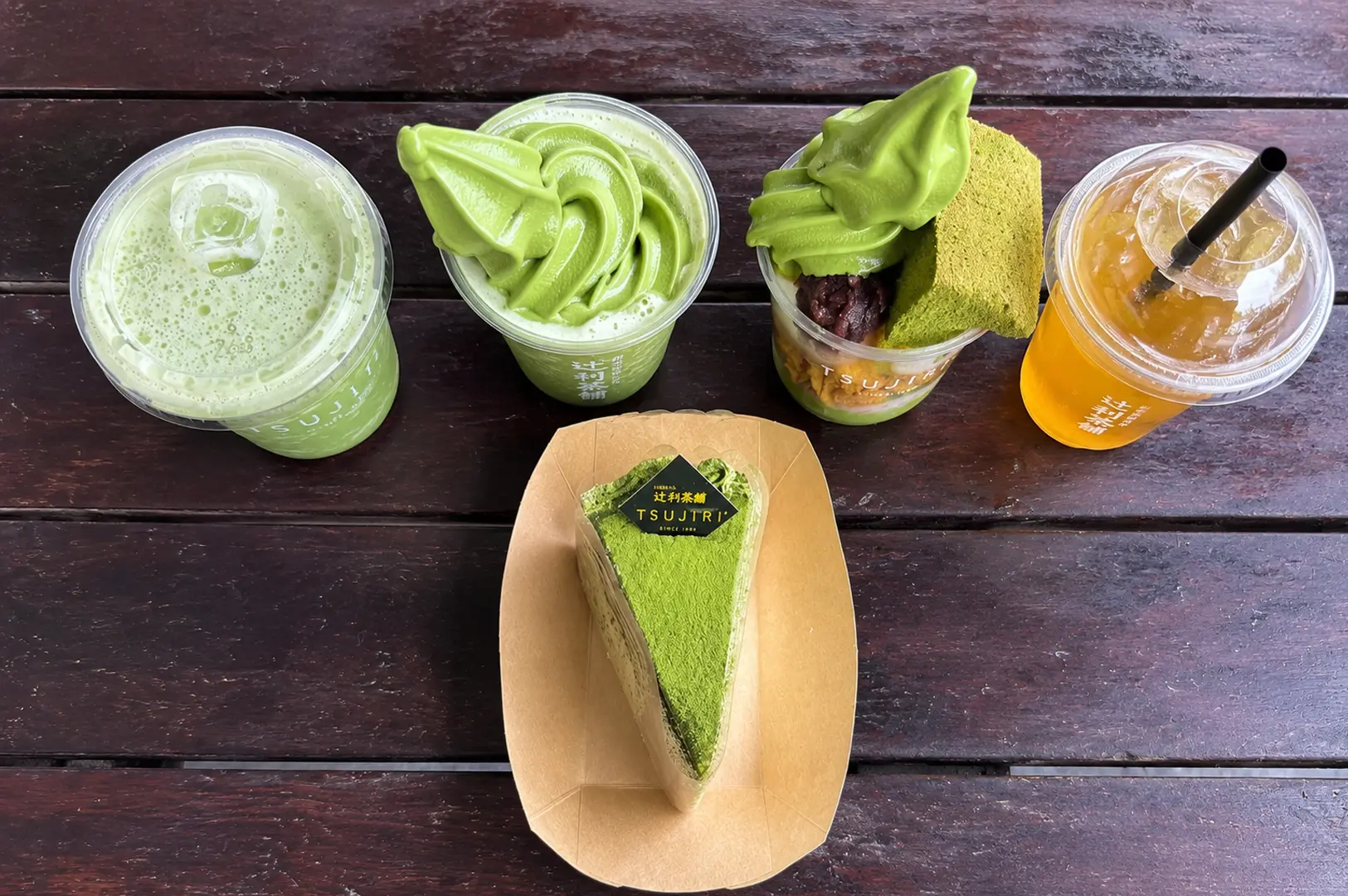 Overhead view of Tsujiri matcha assortment including soft serve, parfait, cake slice, and drinks on wooden table