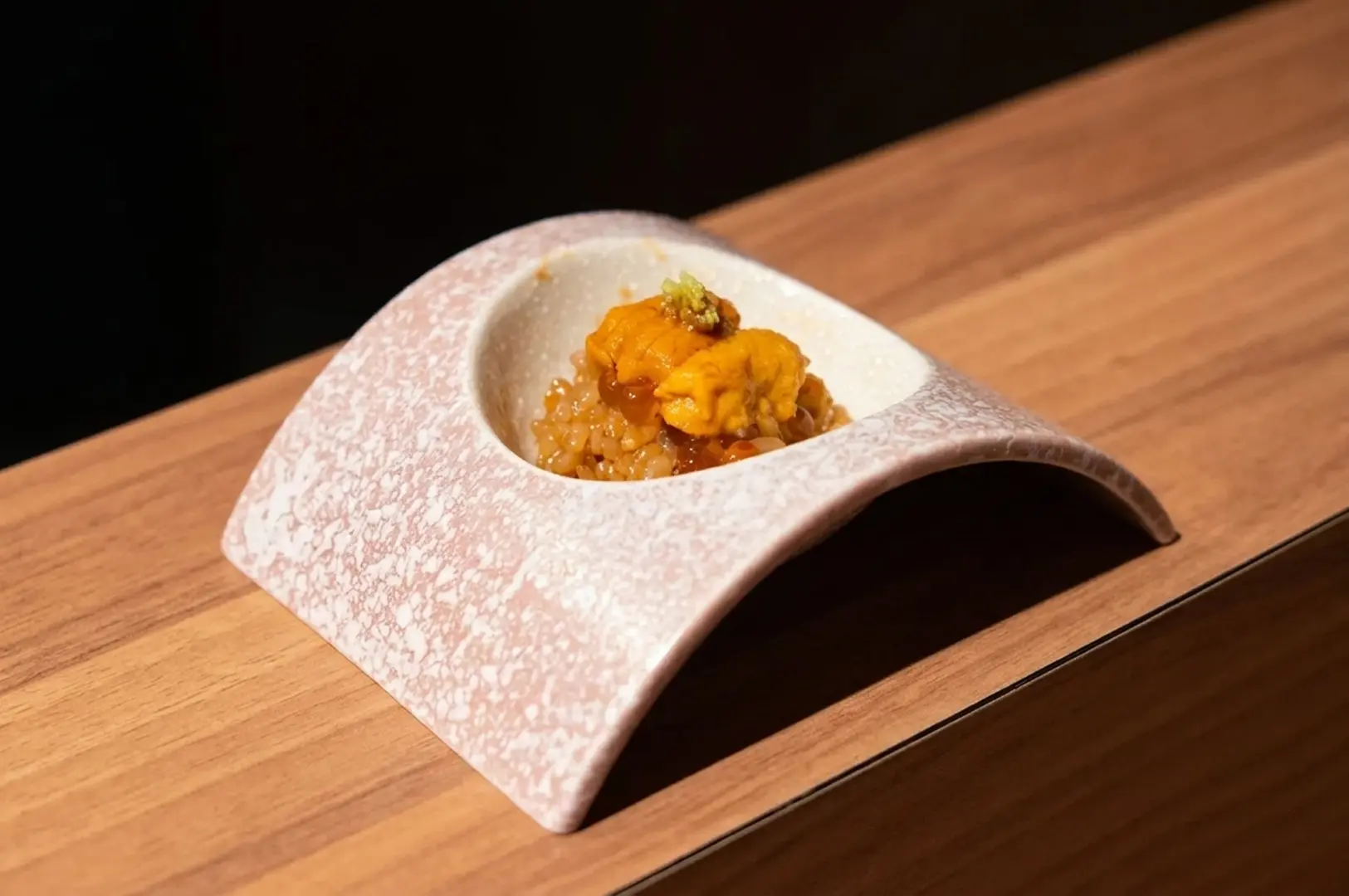 Artistic presentation of seasonal uni and rice served in a unique curved ceramic vessel on a wooden counter