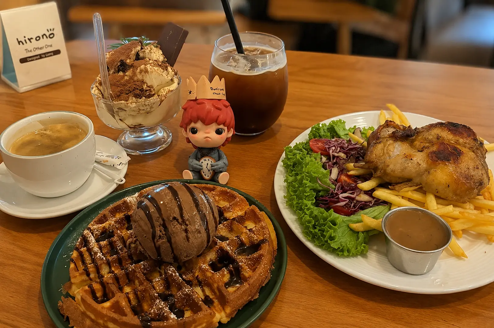 Table spread featuring waffles with ice cream, tiramisu, coffee, and roast chicken with fries at Hirono cafe, accompanied by a small figurine
