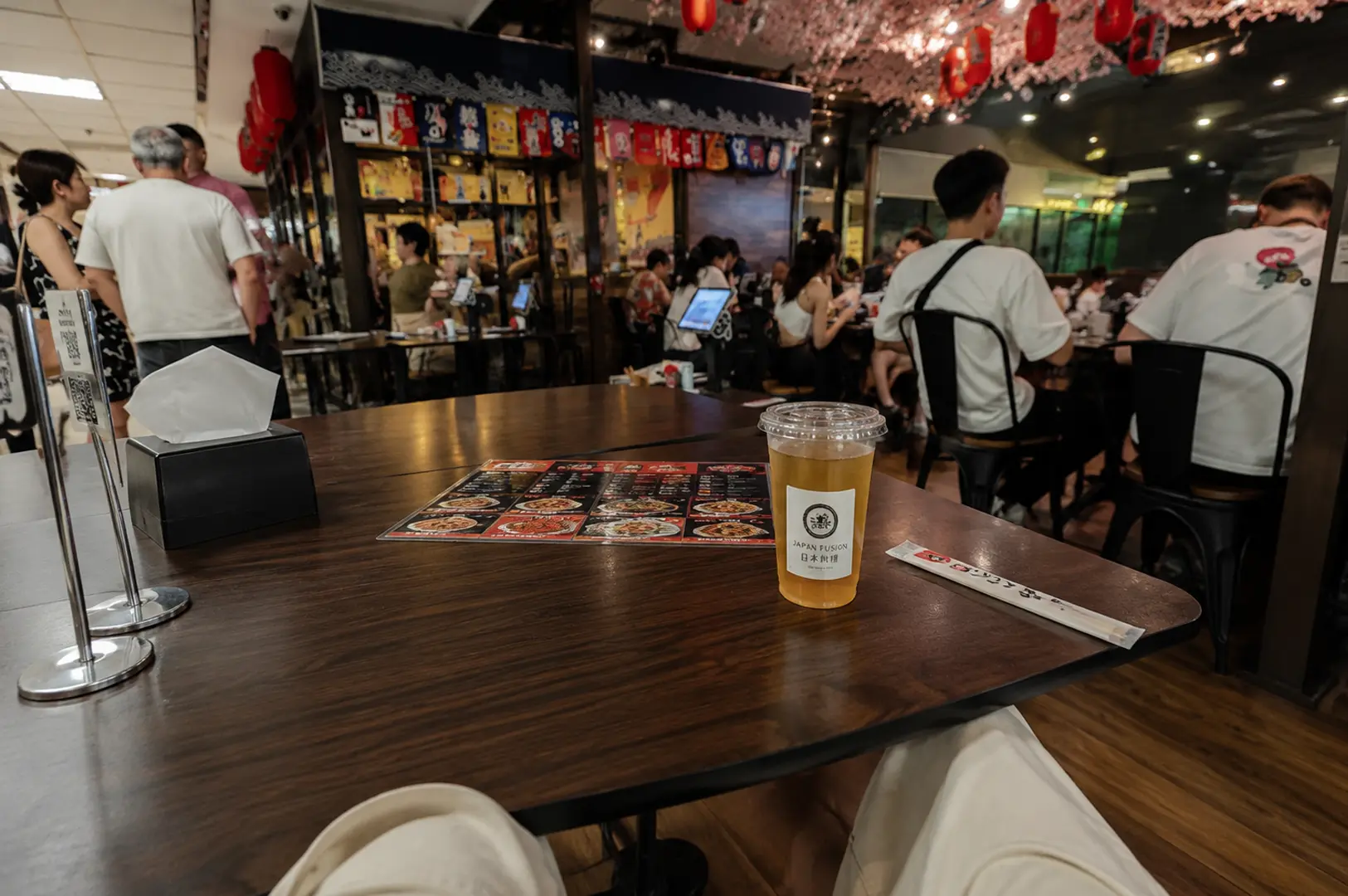 Interior view of a bustling Japanese fusion izakaya with cherry blossom decorations, showing a table set with a menu, drink, and chopsticks