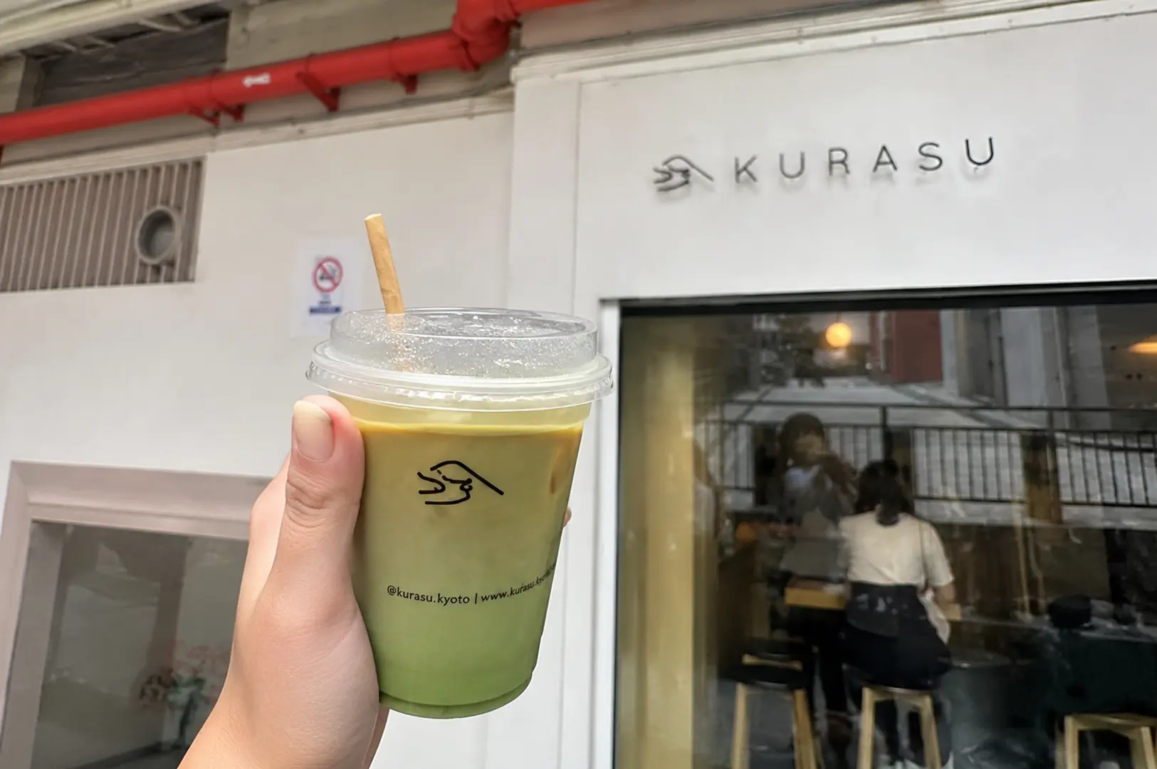 A hand holds an iced matcha latte with the Kurasu logo in front of the KURASU cafe storefront on Waterloo Street.