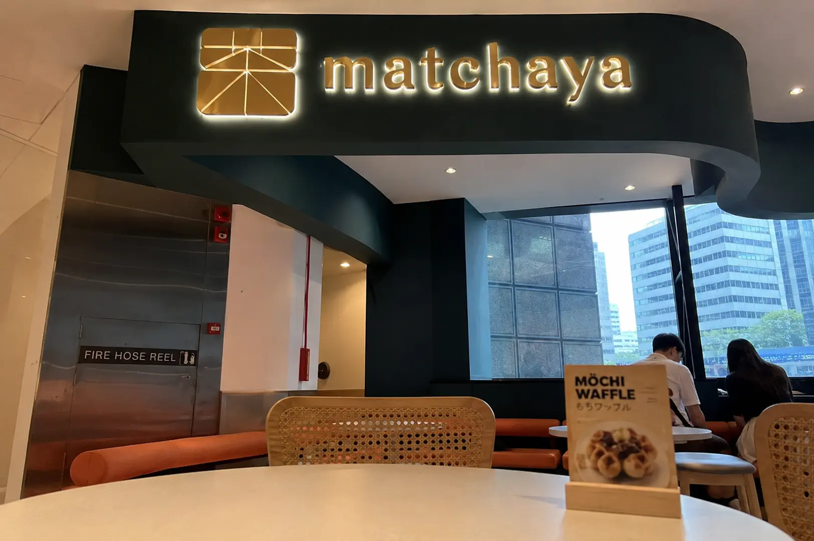 Matchaya cafe interior featuring an illuminated sign, rattan chairs, and a mochi waffle table menu
