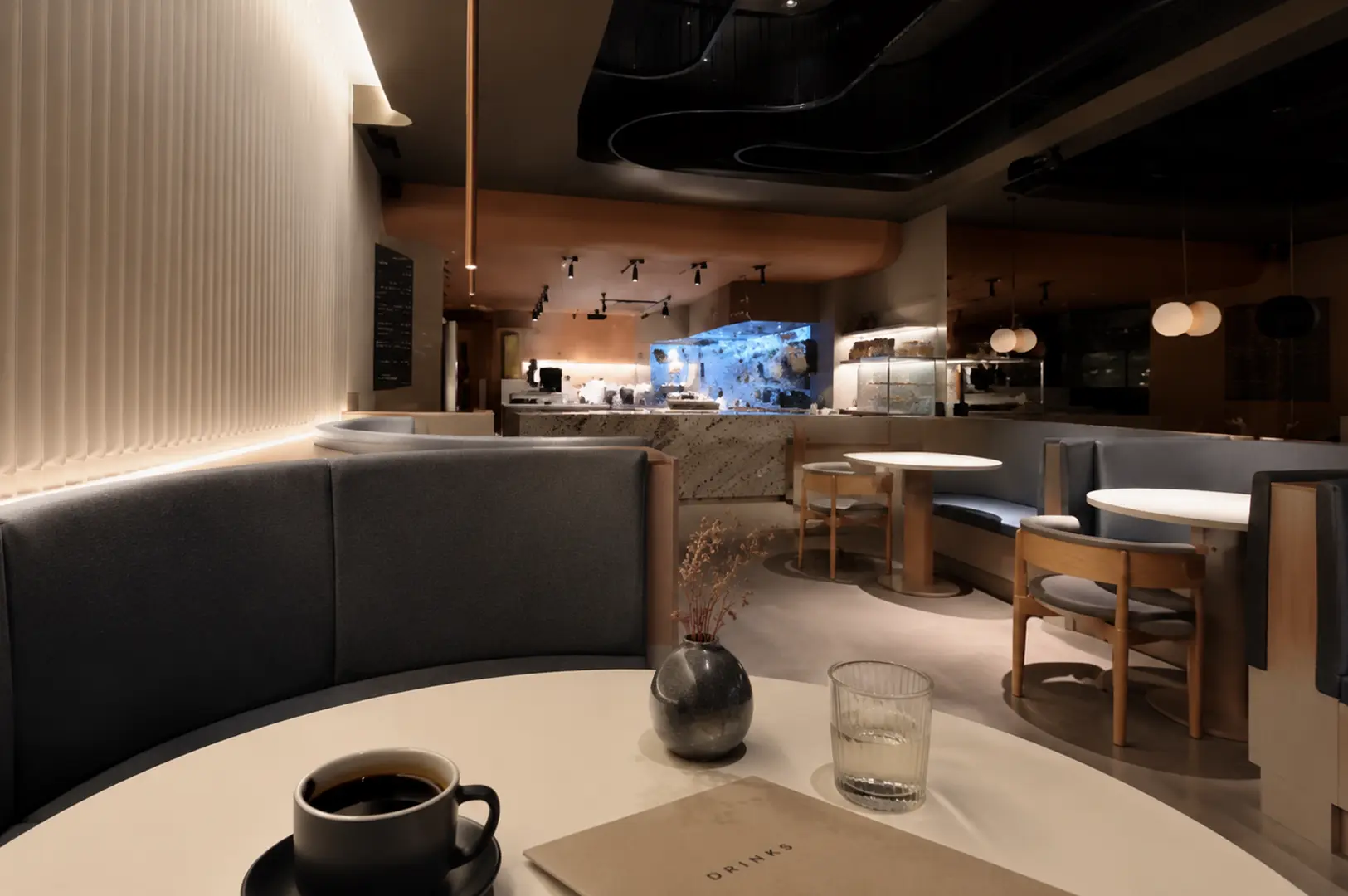 Modern minimalist restaurant interior featuring curved grey booths, white tables, and a blue-lit open kitchen in the background