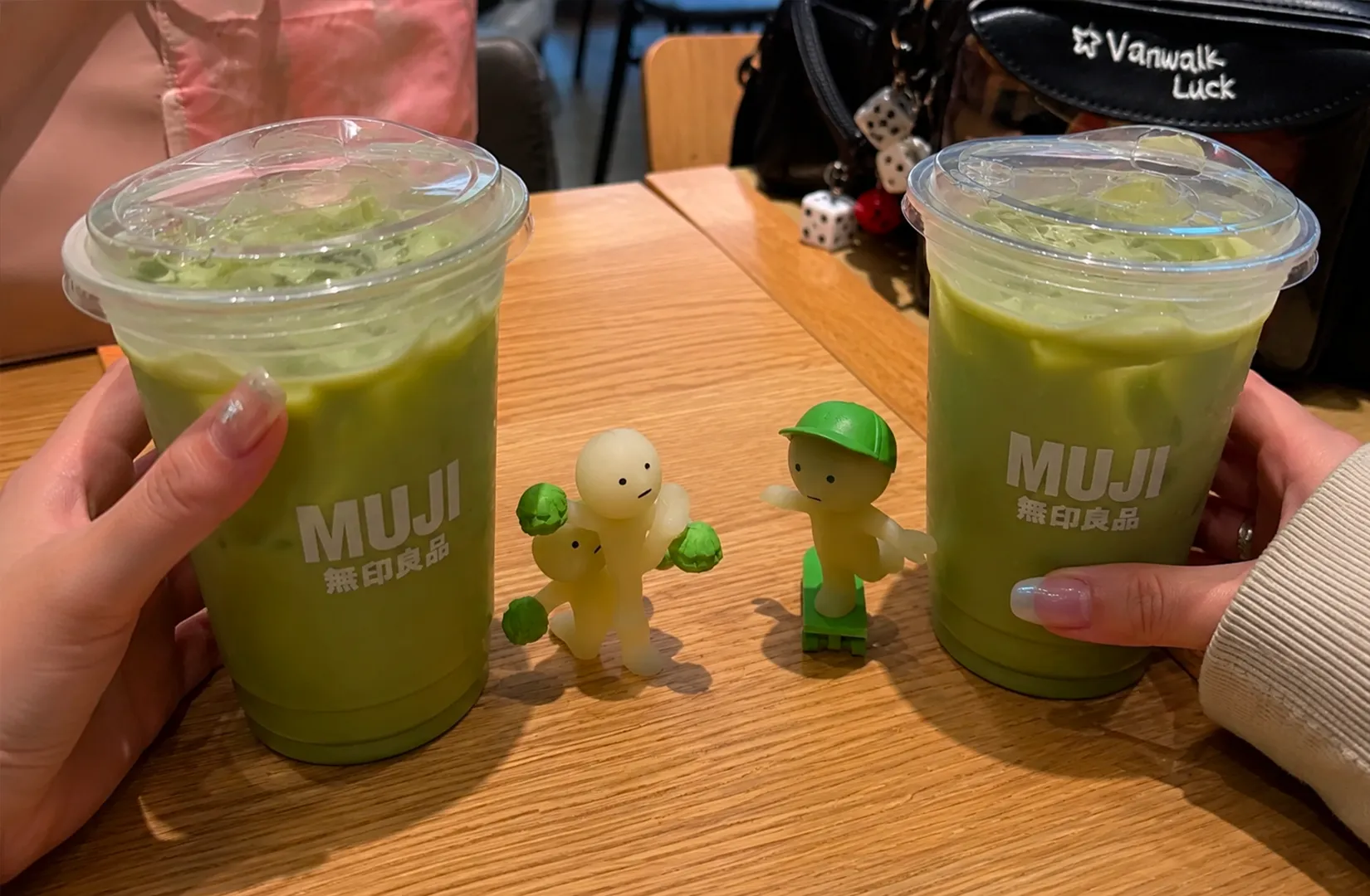 Two hands holding iced matcha lattes in MUJI cups with small figurines on the table
