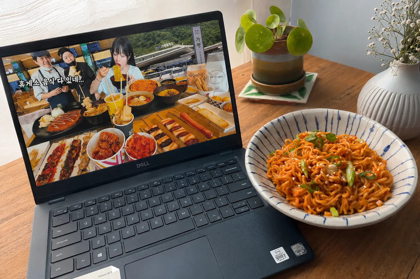 Casual phone photo of a Dell laptop playing a mukbang video beside a bowl of spicy noodles on a wooden table with a small plant and vase