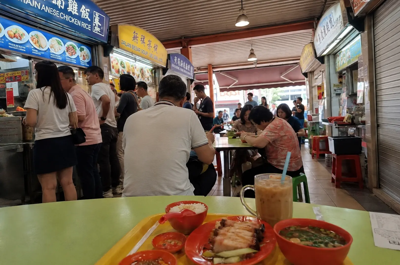 A meal of chicken rice and soup sits on a green table in the foreground, with diners queuing at the Hainanese Chicken Rice and Jalan Besar Soup stalls in the background
