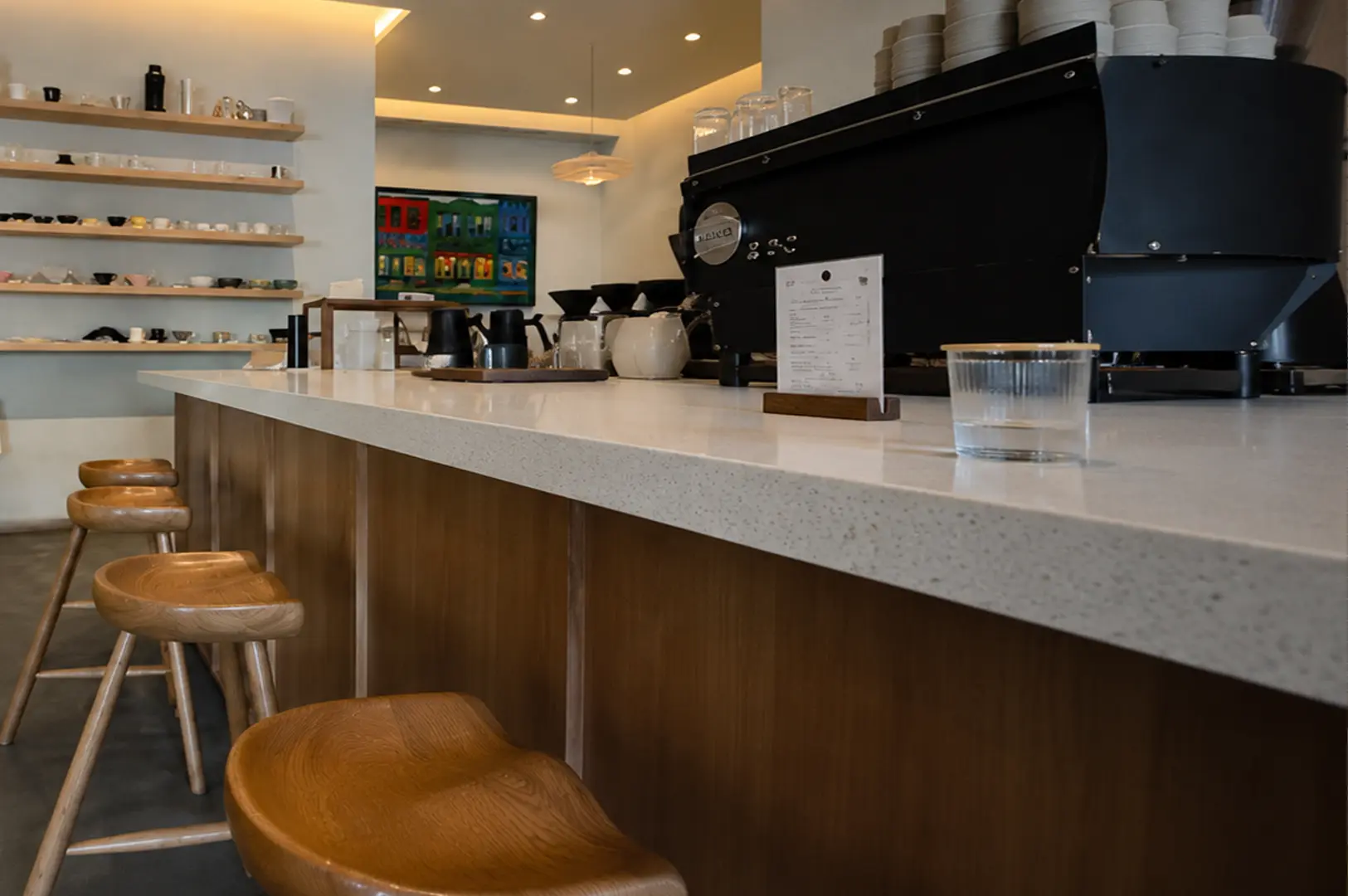 The minimalist cafe interior features a white counter with wooden bar stools, coffee brewing equipment, and open shelving displays