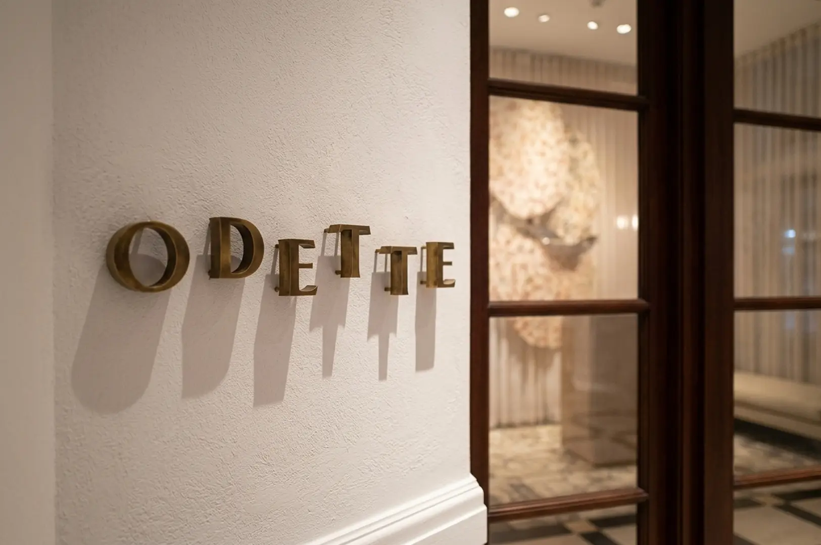 Elegant gold "Odette" lettering mounted on a white textured wall next to a wooden framed glass door